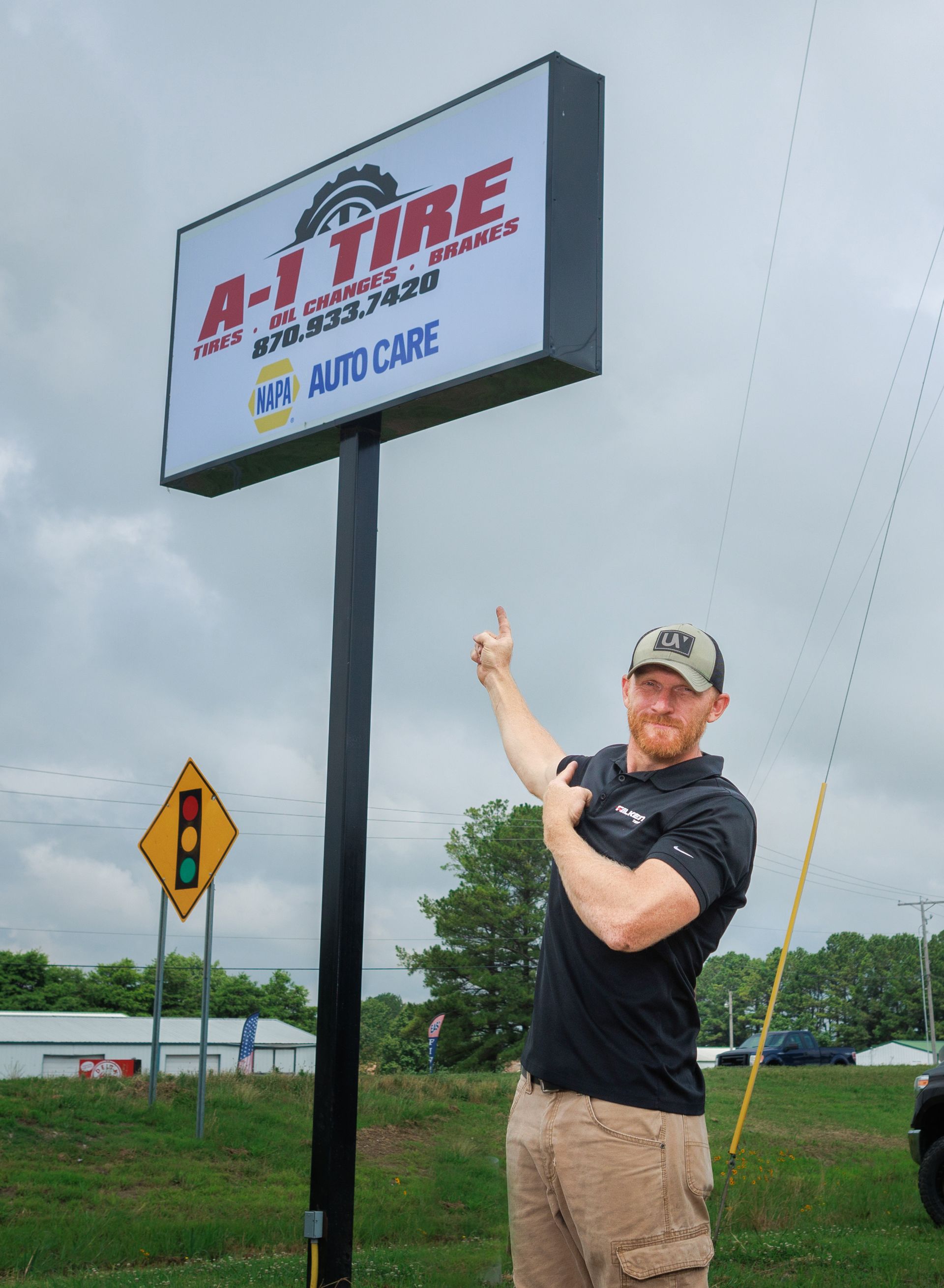Man points to A-1 Tire sign. Sign on black pole, blue and red text. Green grass and cloudy sky.