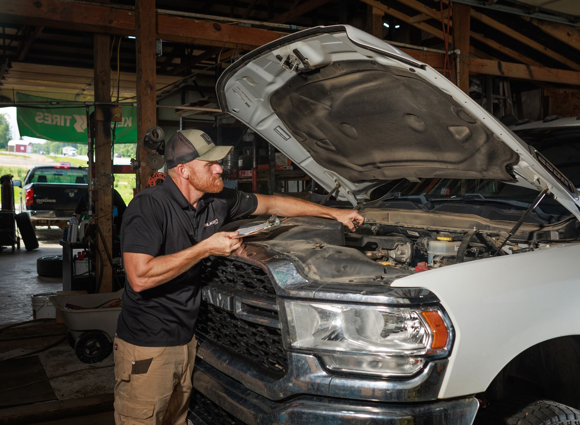Mechanic works on a white truck with open hood in a garage. He wears a hat and black shirt, focused on engine.
