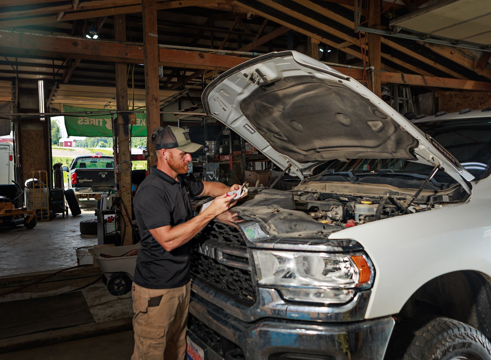 Mechanic inspecting truck engine in a cluttered garage, wearing a cap, holding a tool, and looking at the engine.