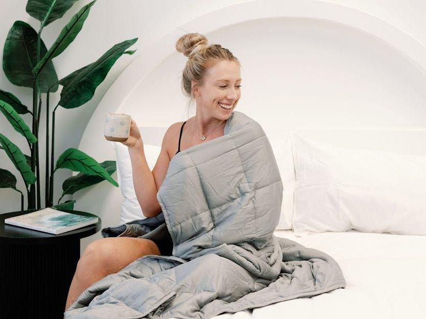 A woman is sitting on a bed wrapped in a blanket holding a cup of coffee.