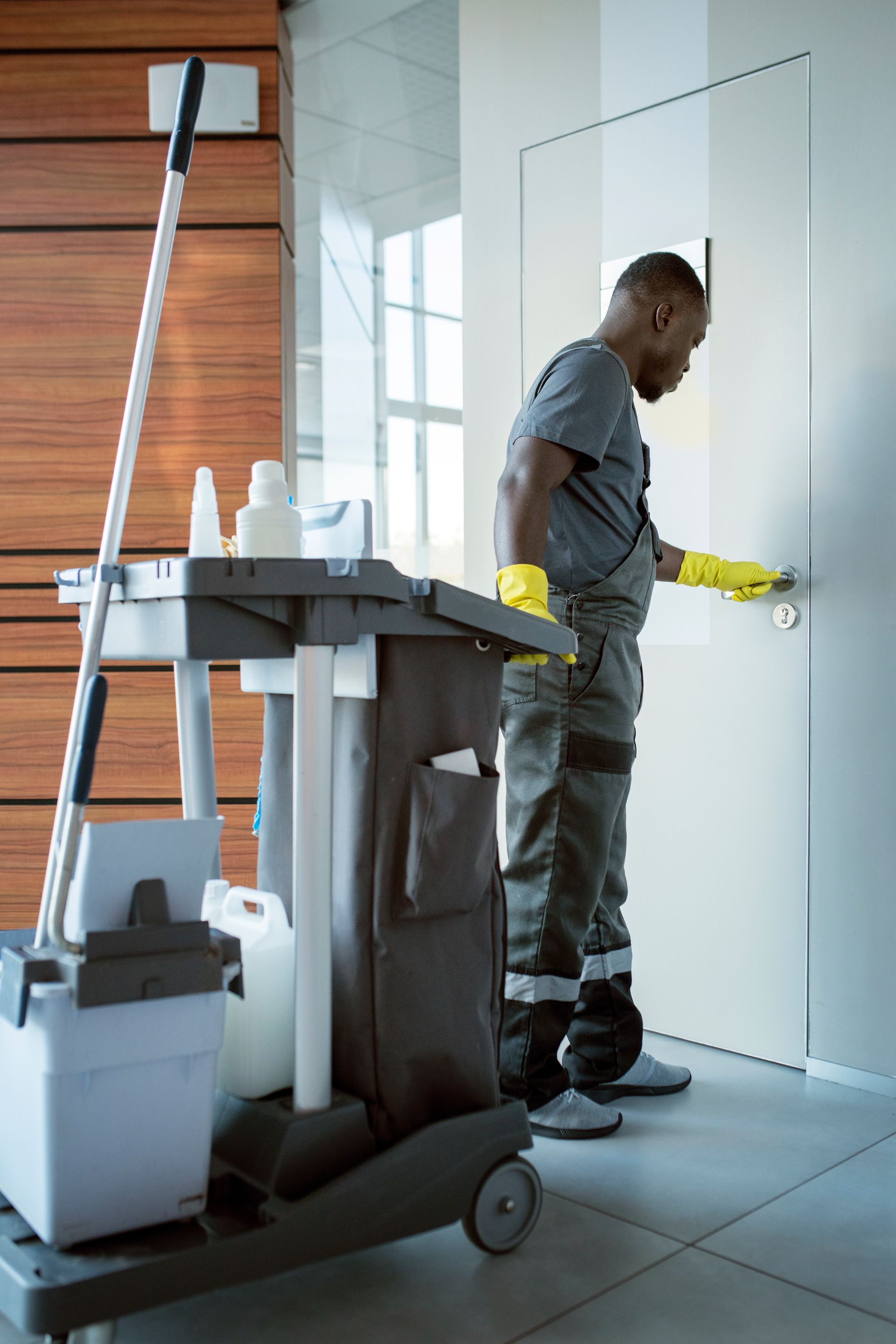 Man in grey uniform and gloves opening door, cleaning cart nearby.