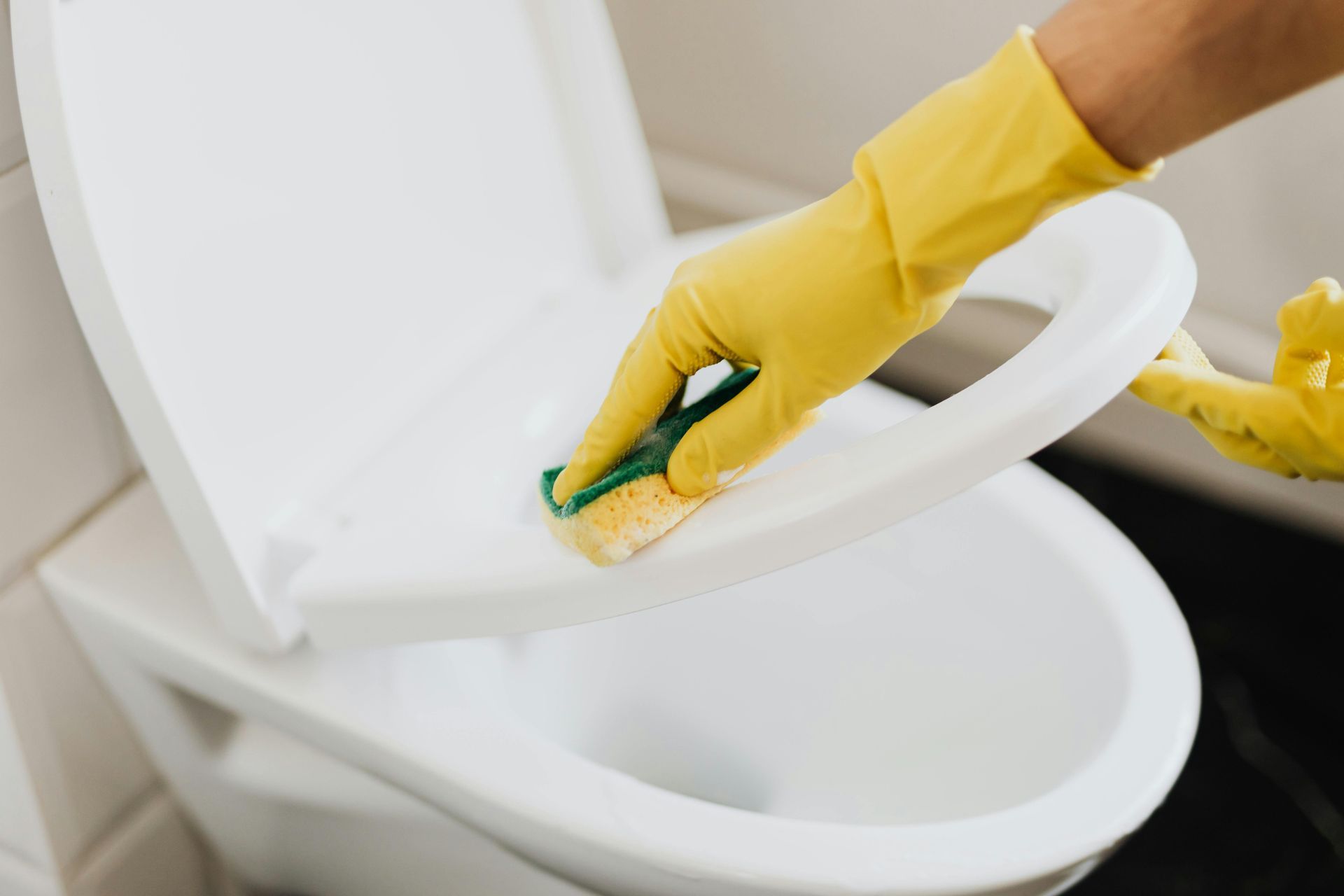 Person wearing yellow gloves cleaning a white toilet seat with a sponge in a bathroom.
