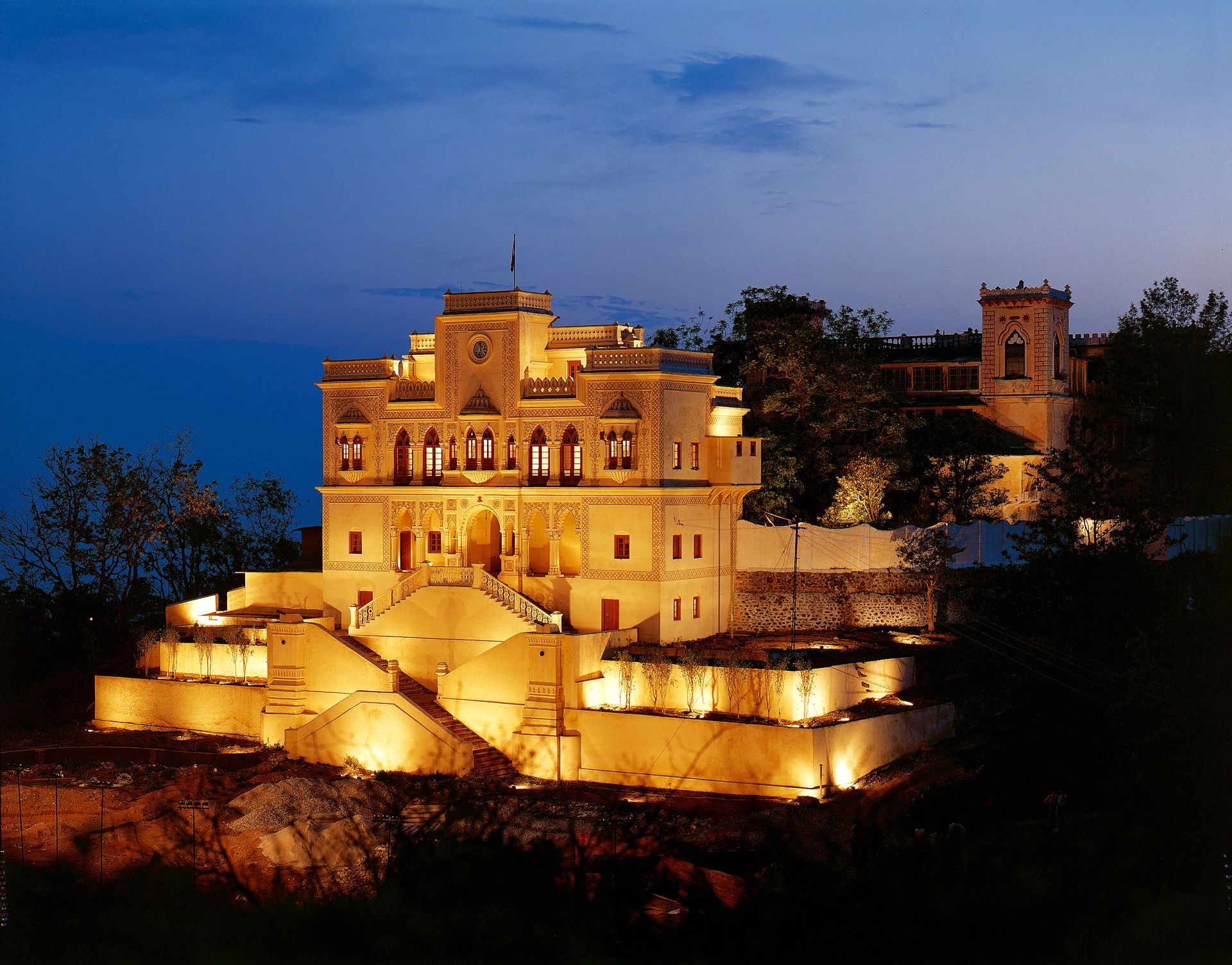 Lit up heritage palace at dusk. Beige building with tiers and arches. Trees in silhouette.