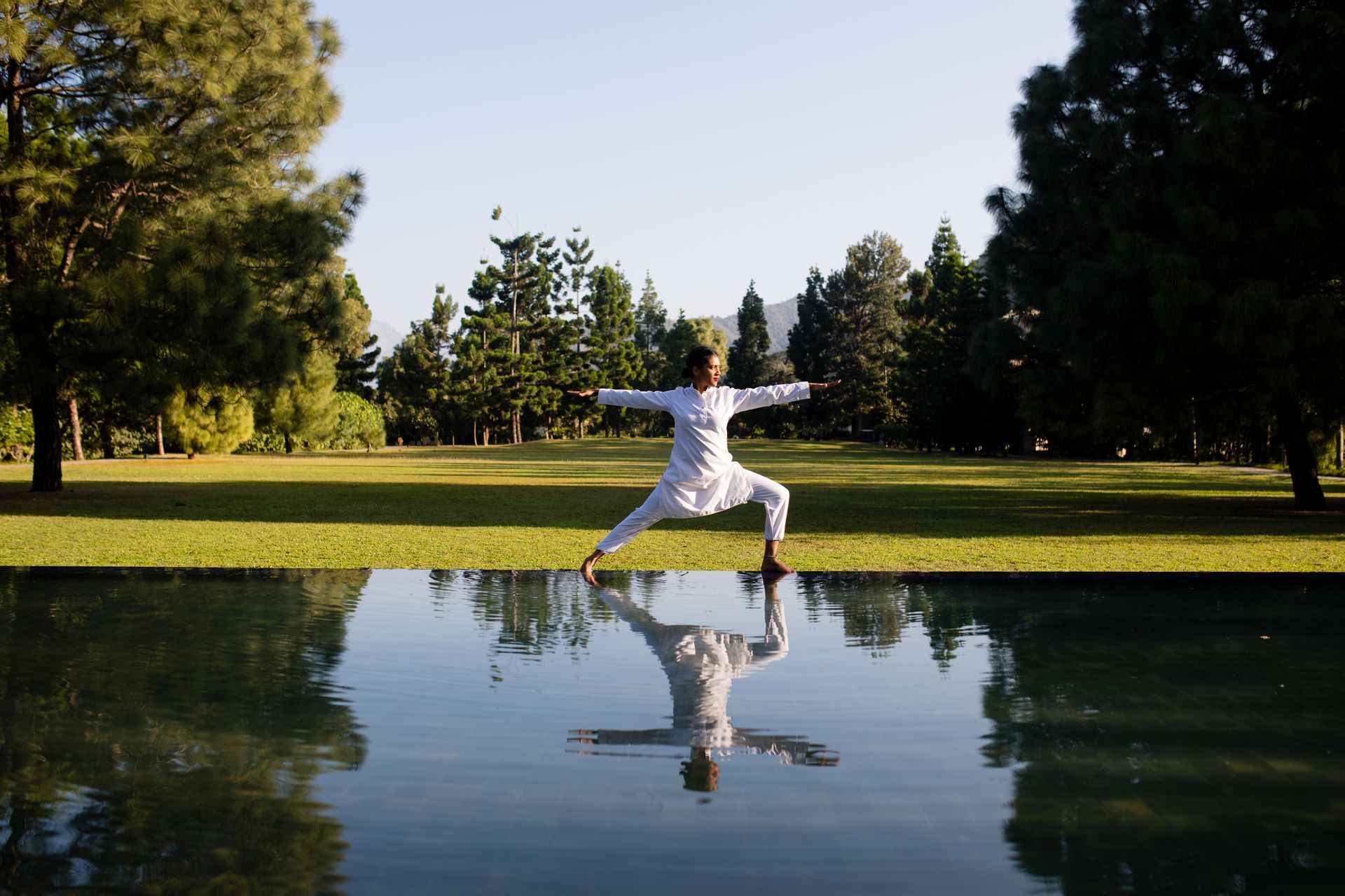 Person in white leaping beside a reflecting pond in a sunny park, with trees lining the background