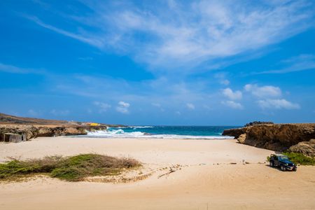 Wide Aruban sandy beach with blue ocean, rocky cliffs, and a small green vehicle under a bright sky