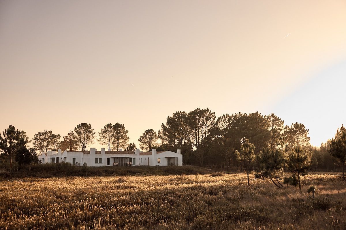 White farmhouse behind a grassy field at sunrise, with trees silhouetted in warm light.