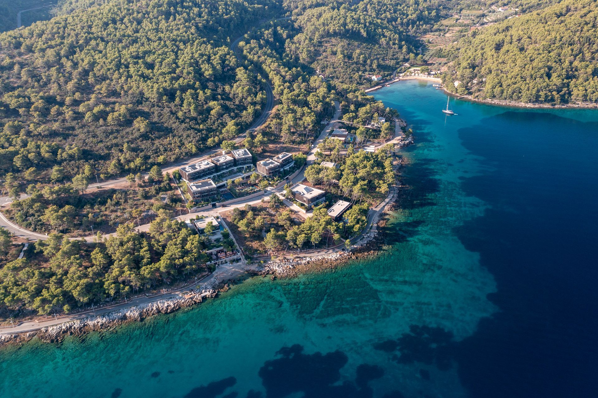 Aerial view of a coastal area with turquoise water, a treed hillside, and buildings.
