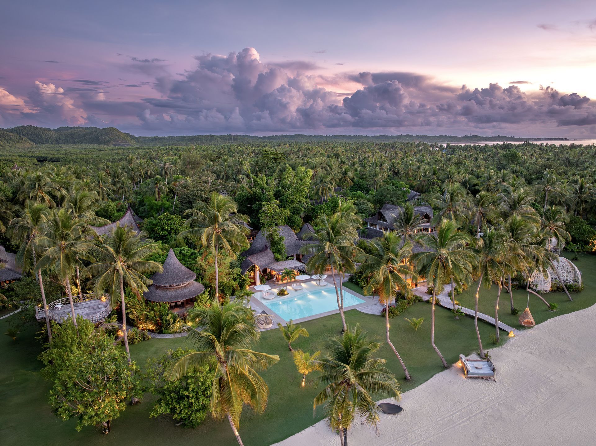 Luxury resort nestled among lush palm trees and green vegetation beside a white sand beach at sunset.