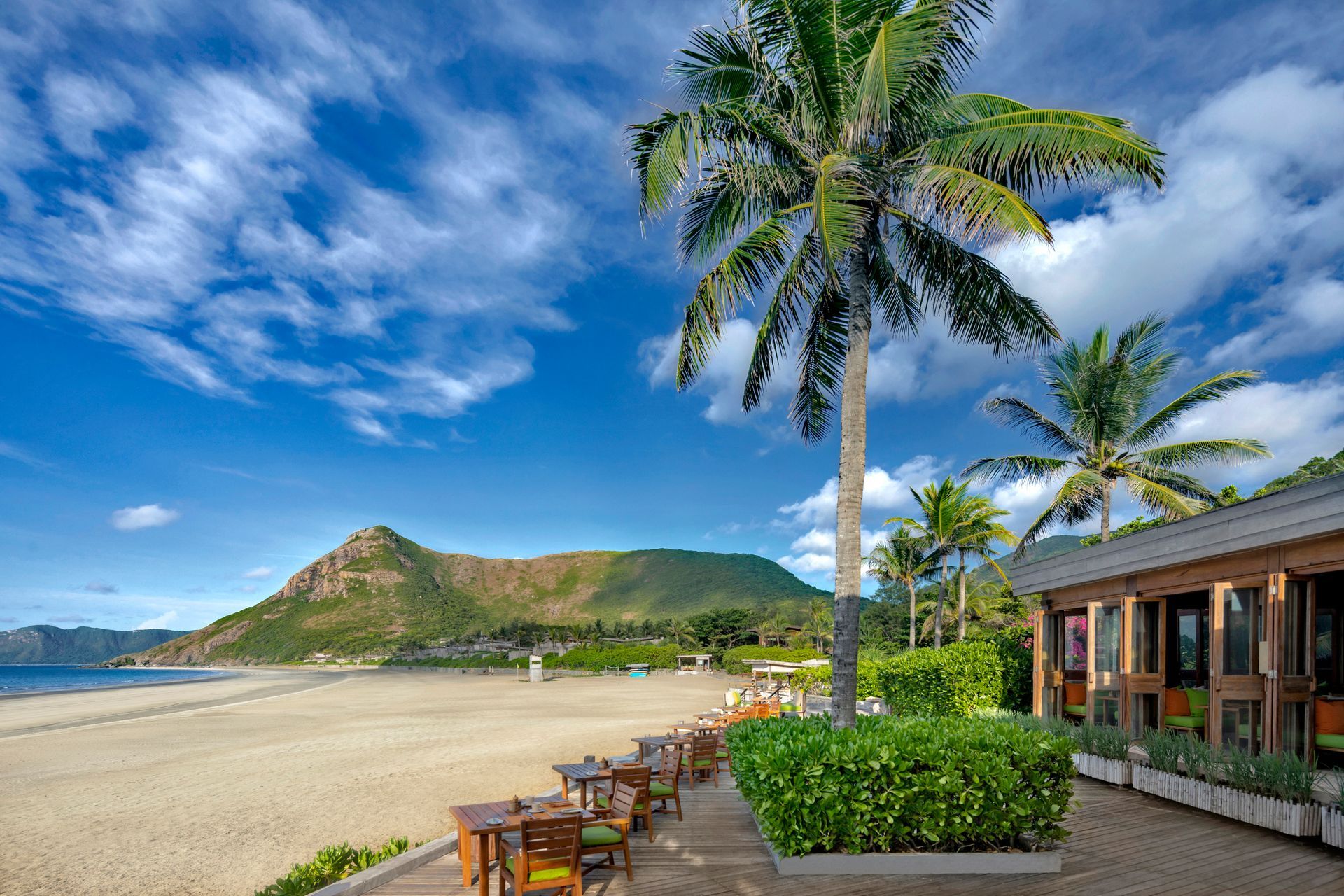 Beach scene with palm trees, sandy shore, restaurant patio, and mountain under a blue, cloudy sky.