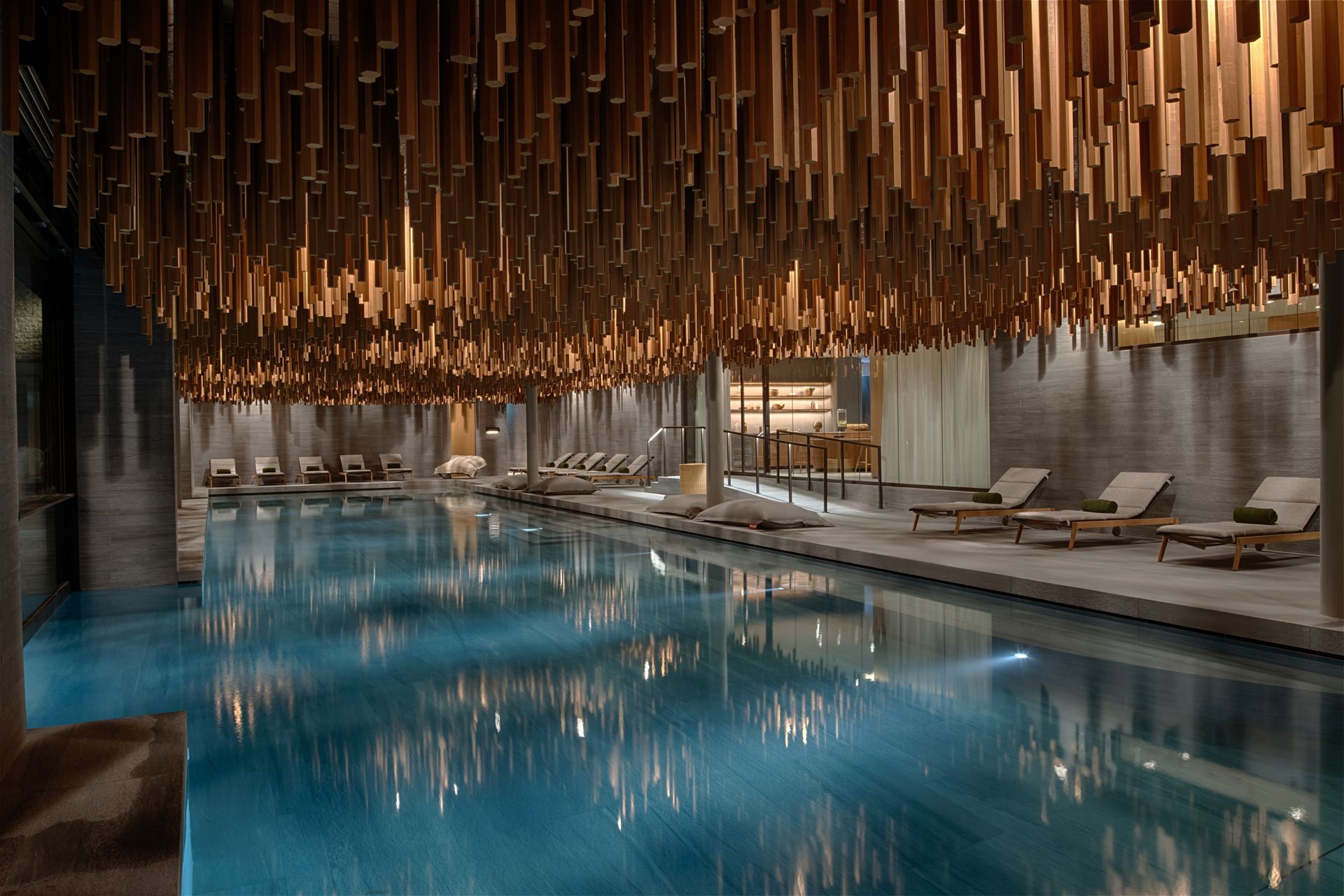 Indoor pool with wood slat ceiling; lounge chairs line the pool's side. Blue water reflects light.