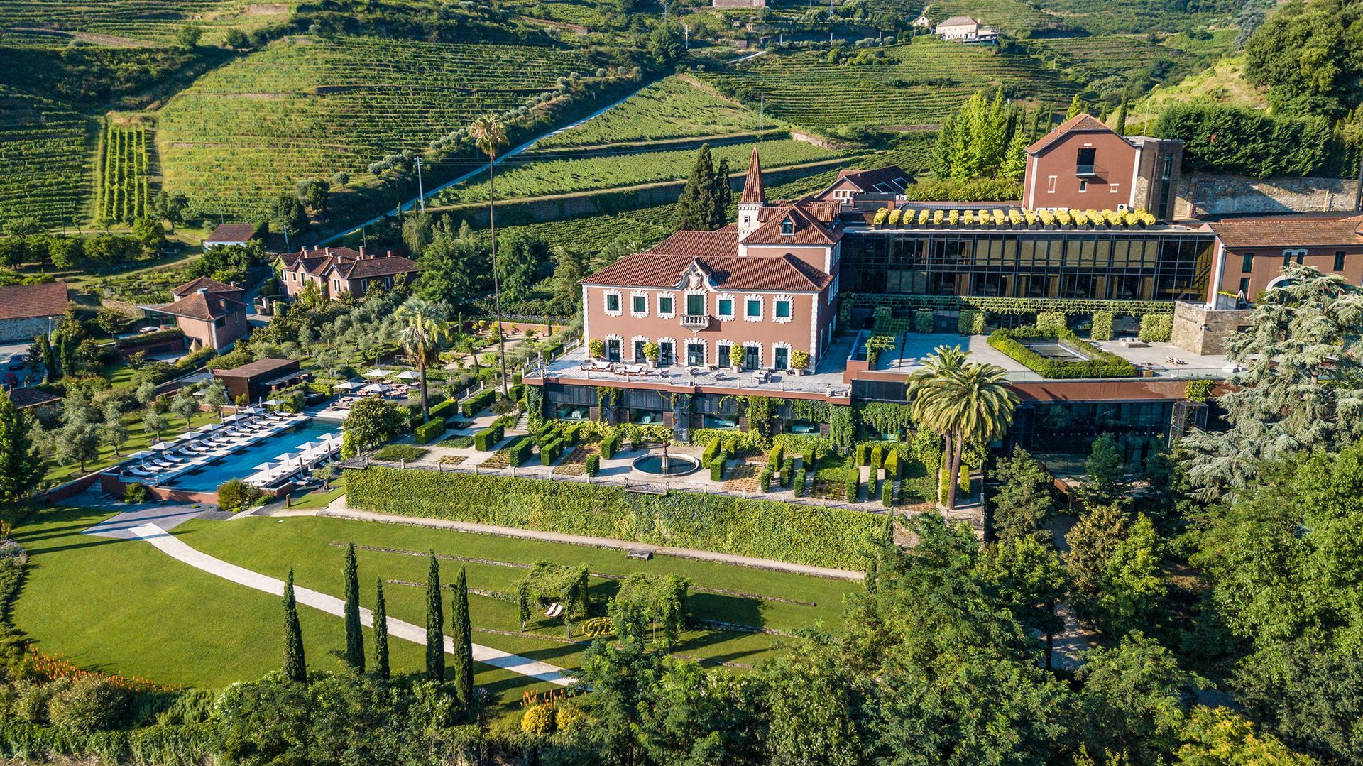 Aerial view of a villa with a pool, gardens, and vineyards in a sunny, green landscape.