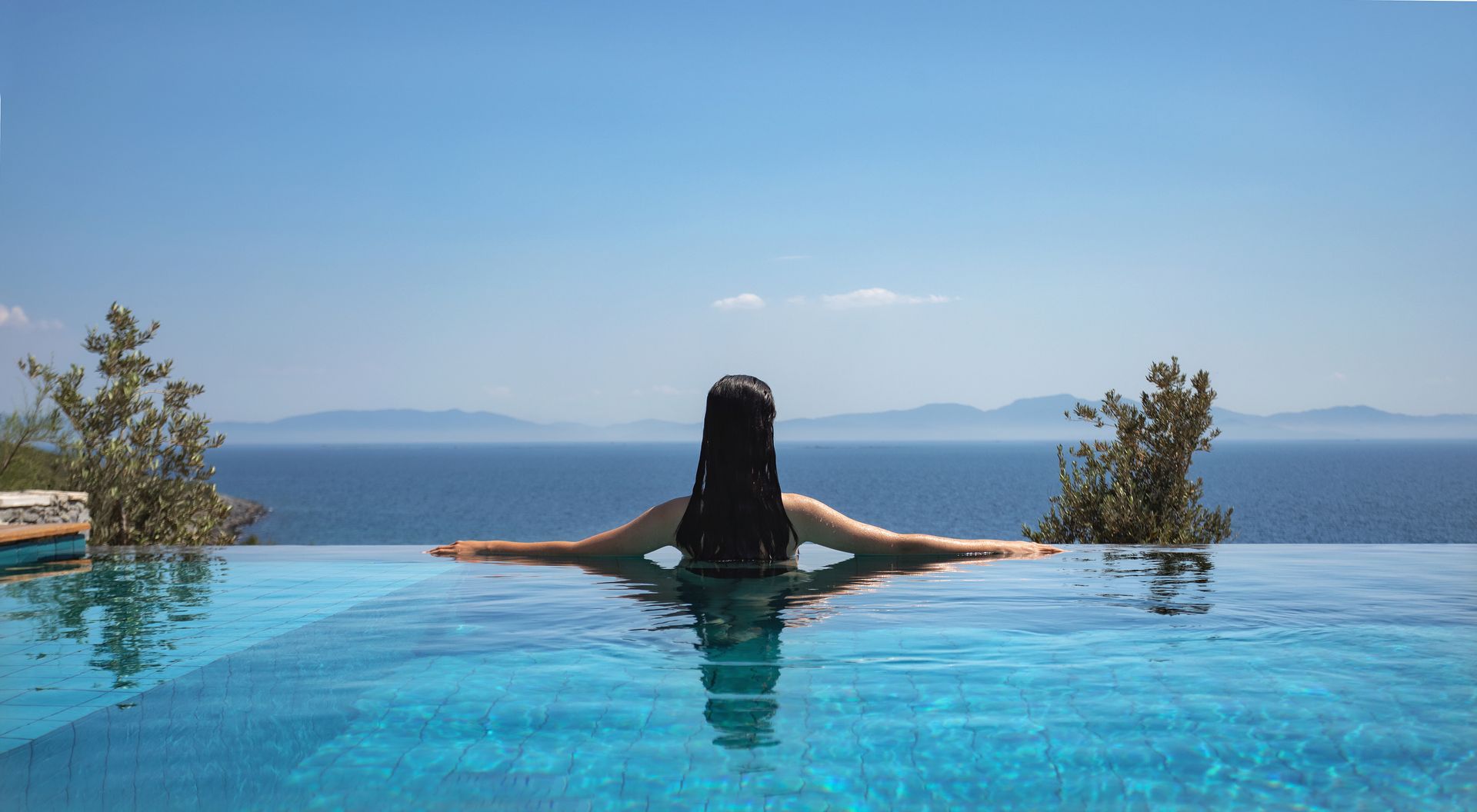 A woman is sitting in an infinity pool overlooking the ocean.