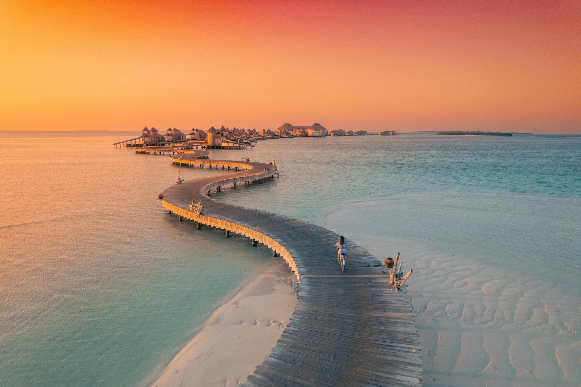 Curved wooden boardwalk over turquoise water at sunset, leading to a small overwater island resort