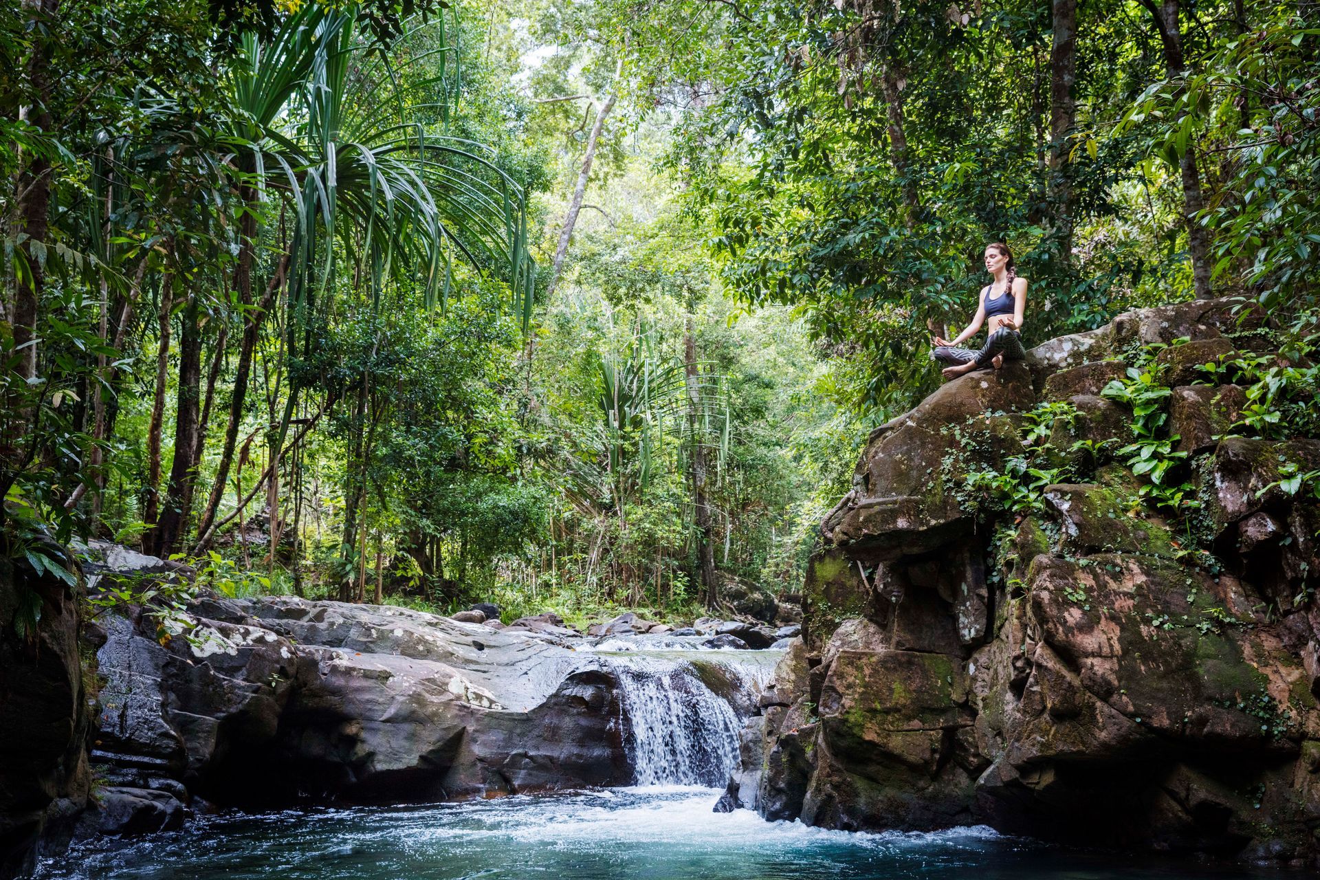 Person sitting on a rock above a small waterfall in a lush green forest at the Datai.