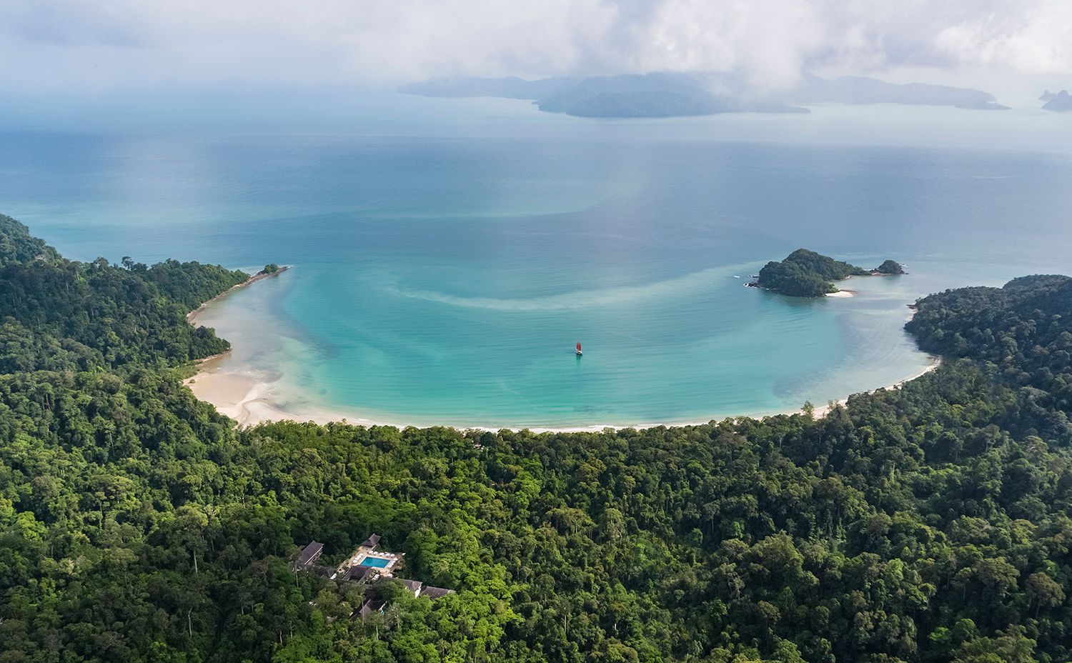 Aerial view of a secluded turquoise bay, white sandy beach, and lush green jungle, under a cloudy sky.