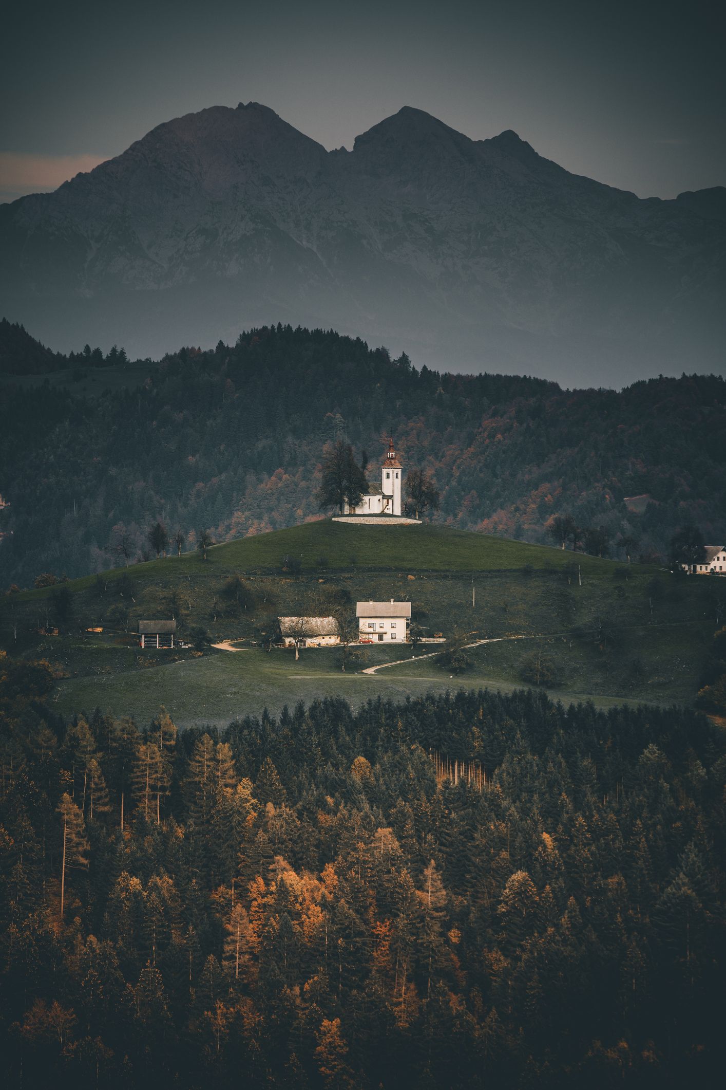 A church on top of a hill in the middle of a forest with mountains in the background.