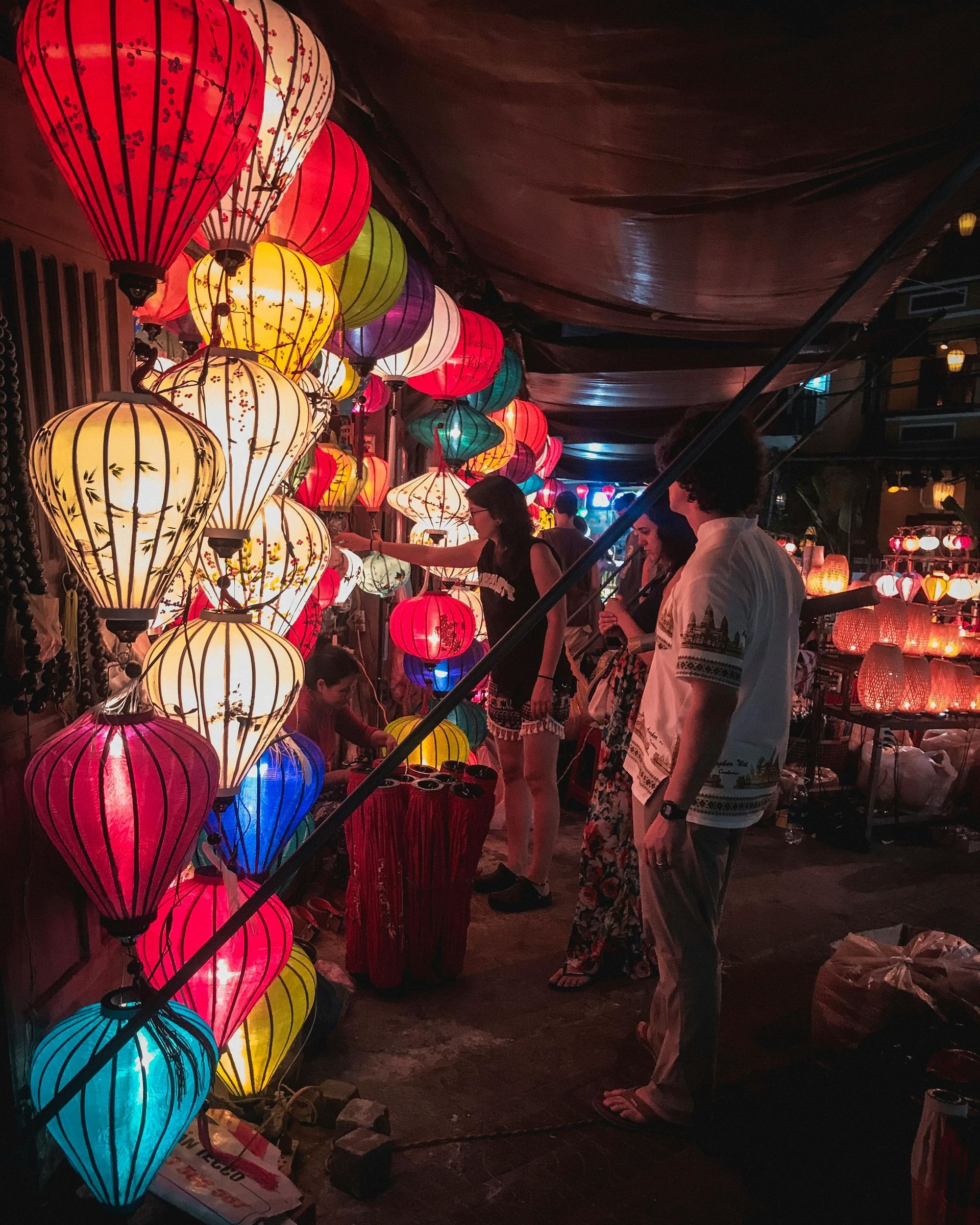 Colorful lanterns illuminate a shop. People browse the lanterns in a dimly lit market.