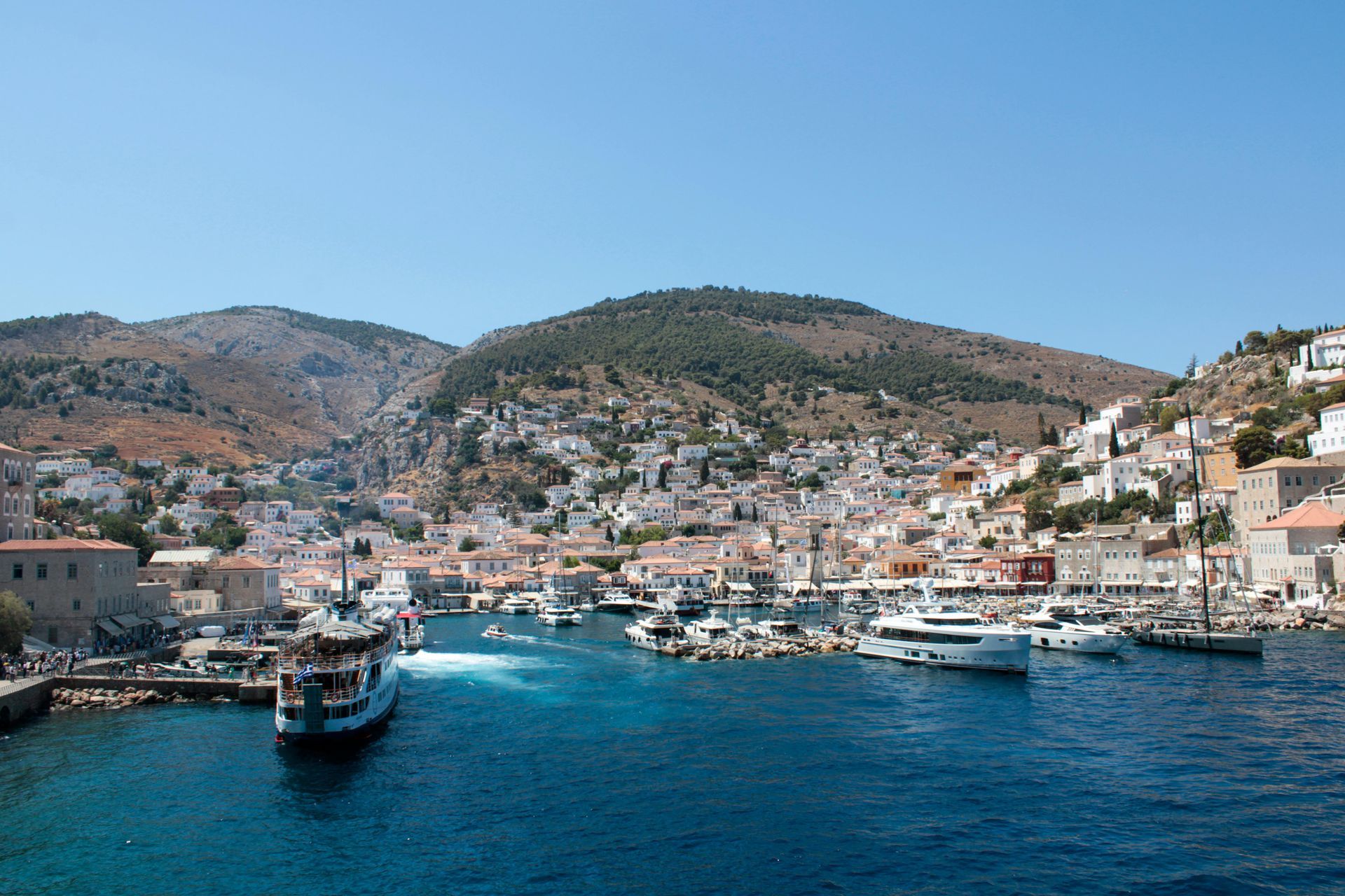 A boat is going through a Hydra harbor with mountains in the background.