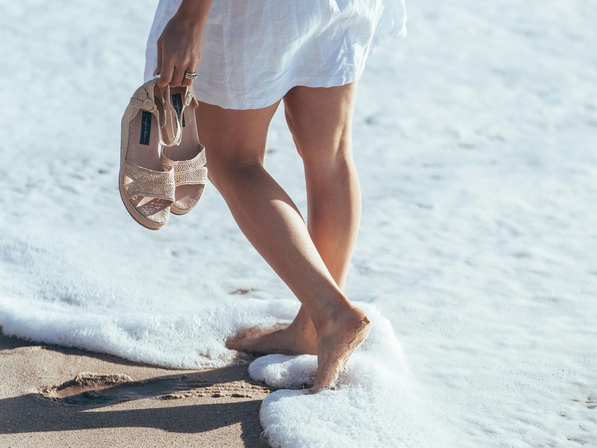 A woman in a white dress is walking on the beach holding a pair of sandals.