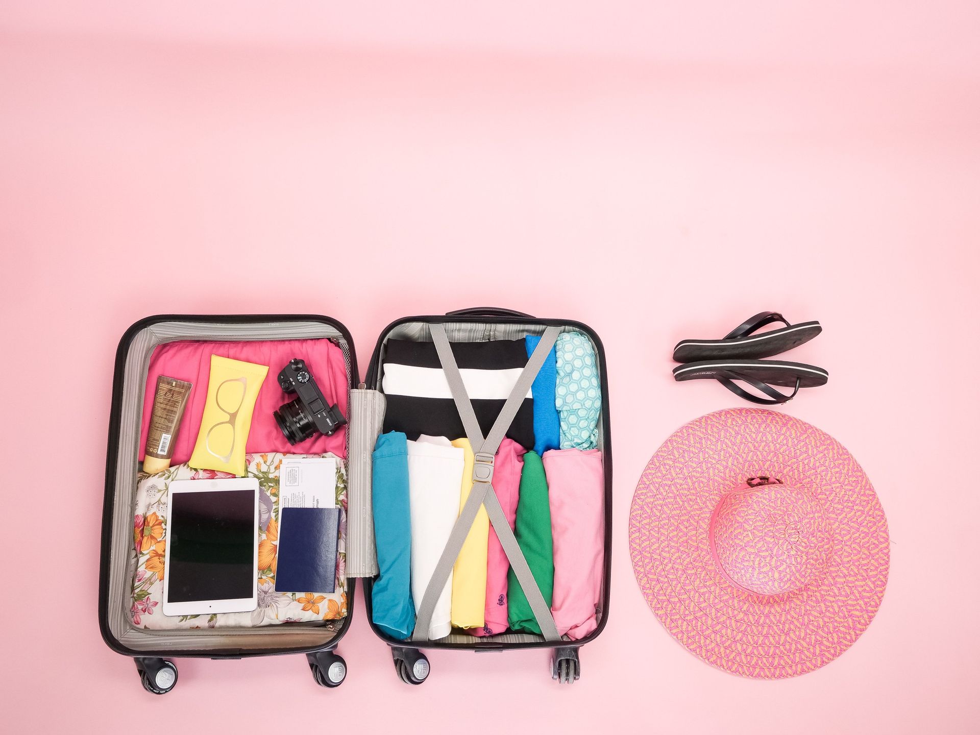 Open suitcase with colorful travel items beside a pink sun hat and sunglasses on a pink background