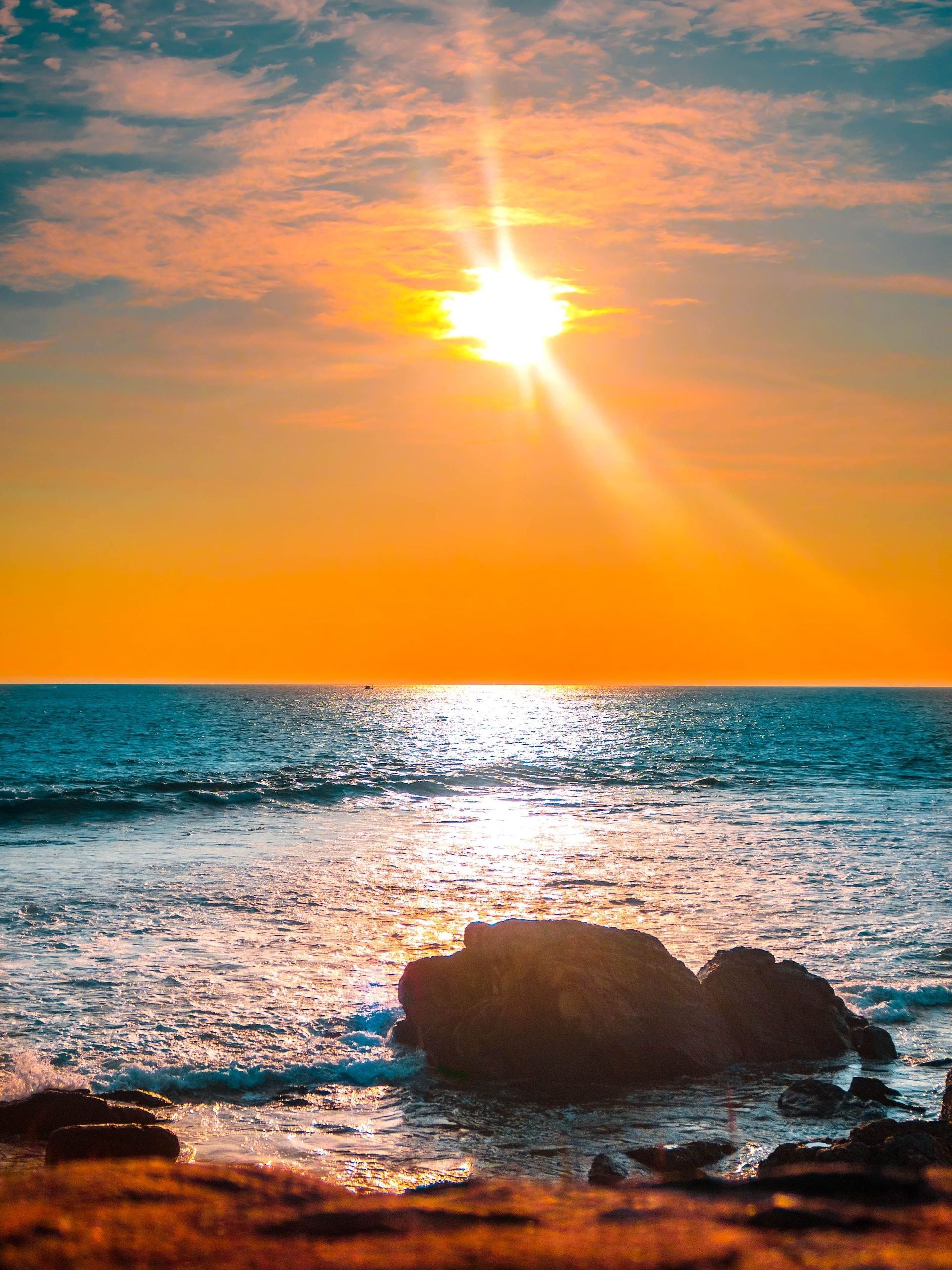 The sun is setting over the ocean with a rock in the foreground.