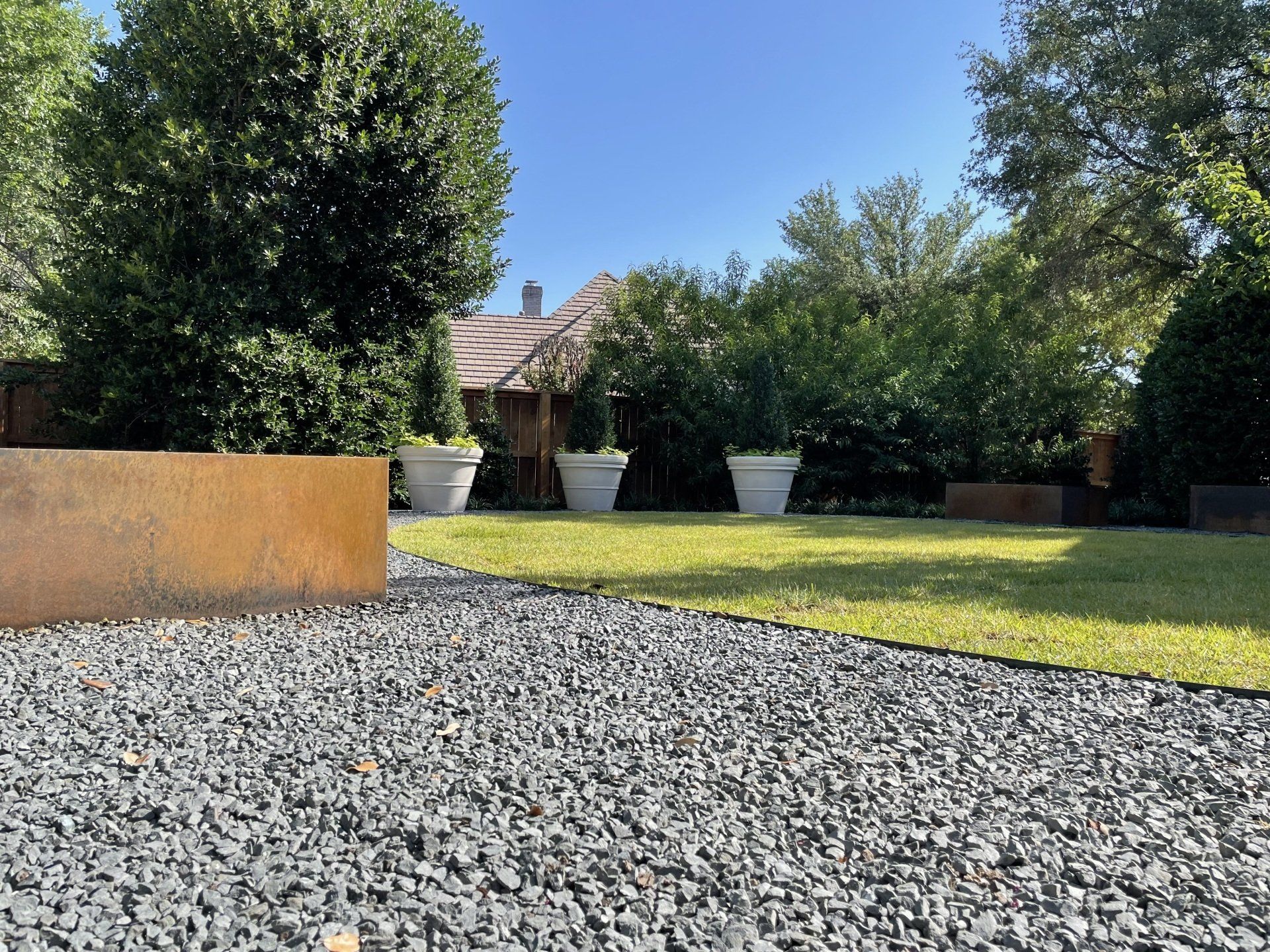 Gravel path leads to lawn with planters, trees, and a house on a sunny day.
