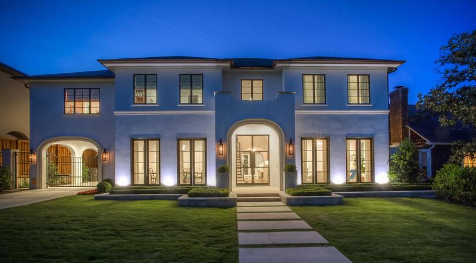 Two-story blue house at dusk, lit up by exterior lights, with a green lawn and a path leading to the front door.