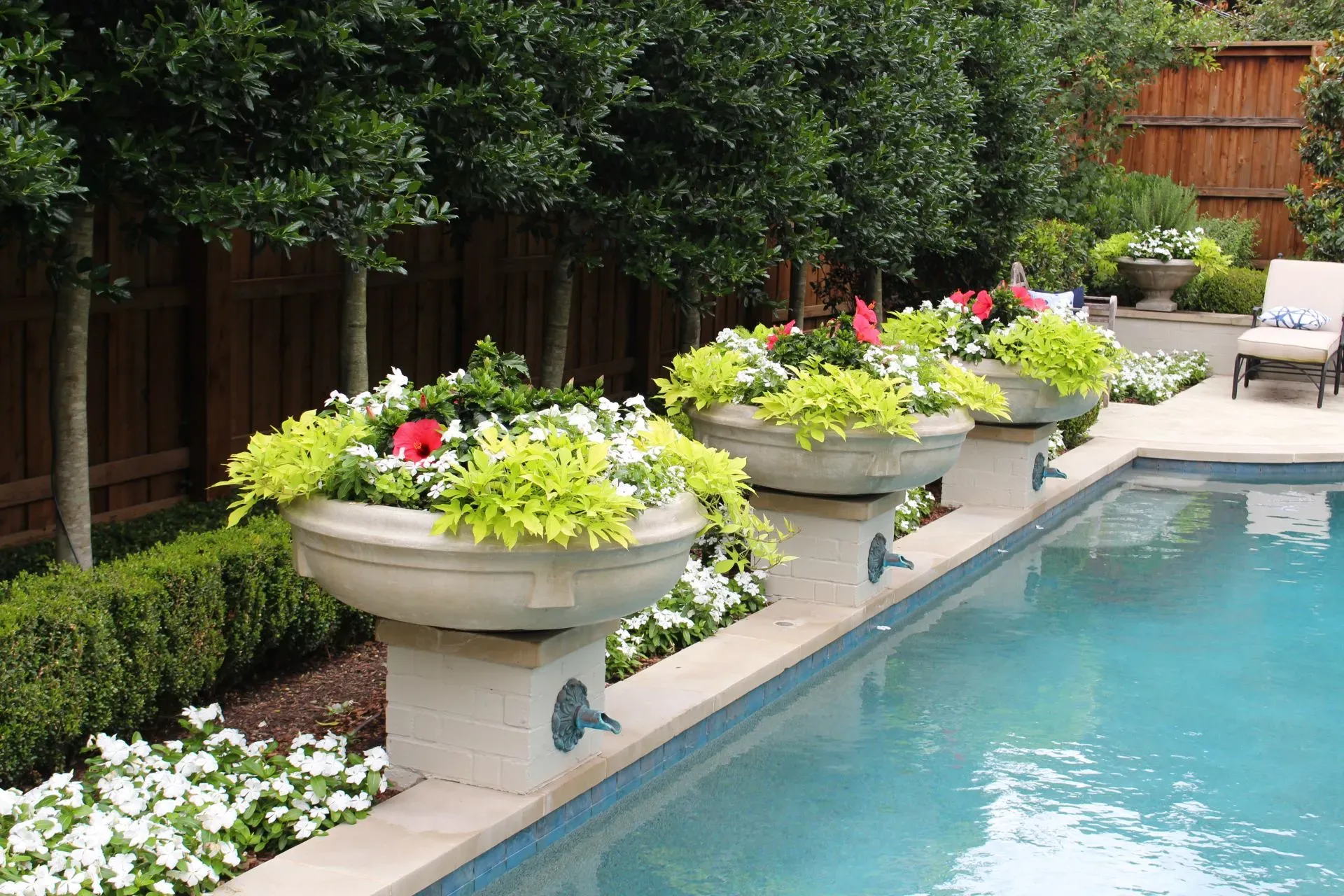 Poolside garden with flower-filled planters, bordering a pool with clear water. 