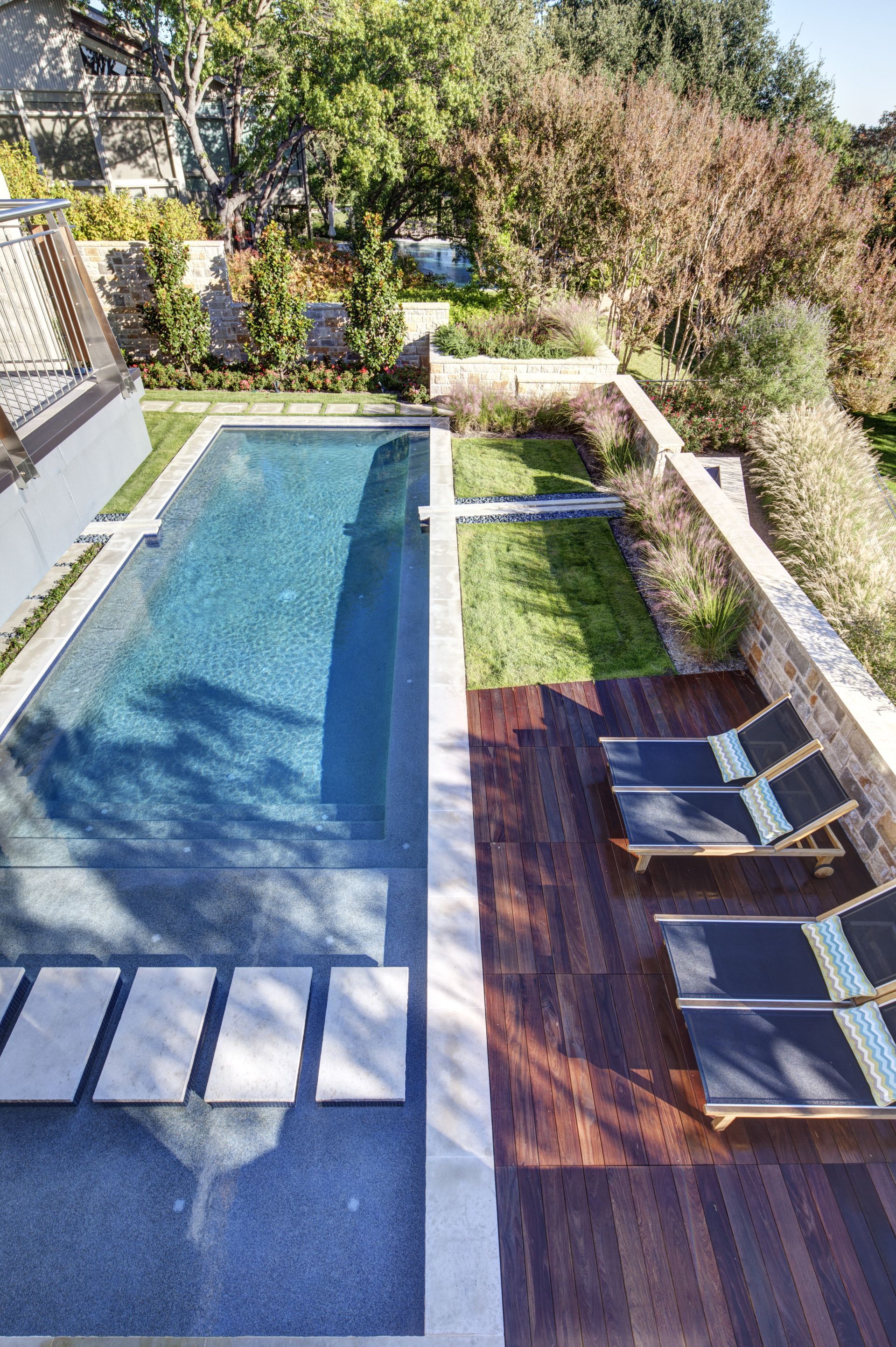 Long rectangular pool with wooden deck and lounge chairs; greenery and stone wall surround.