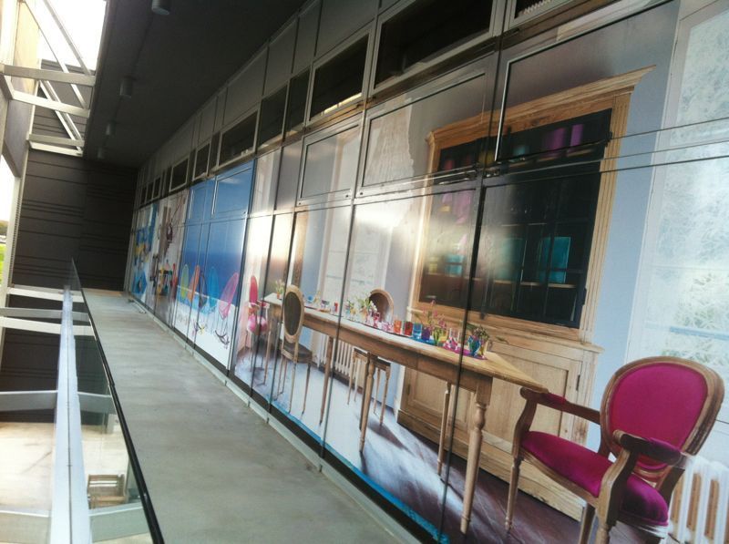 Lungo corridoio con grandi immagini stampate di mobili, un tavolo e una sedia rossa sui pannelli di vetro.