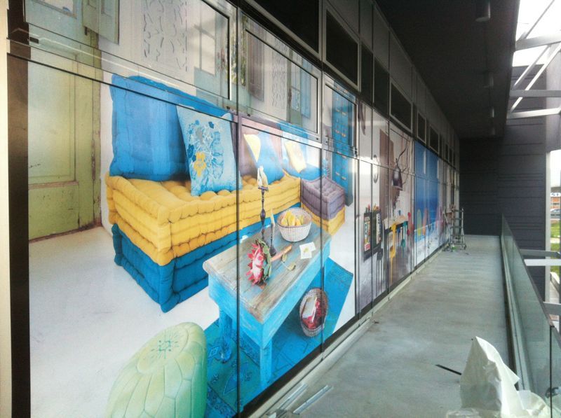 Grande murale raffigurante una scena d'interni dai colori vivaci, con un divano blu, cuscini gialli e un tavolino da caffè su un passaggio esterno.