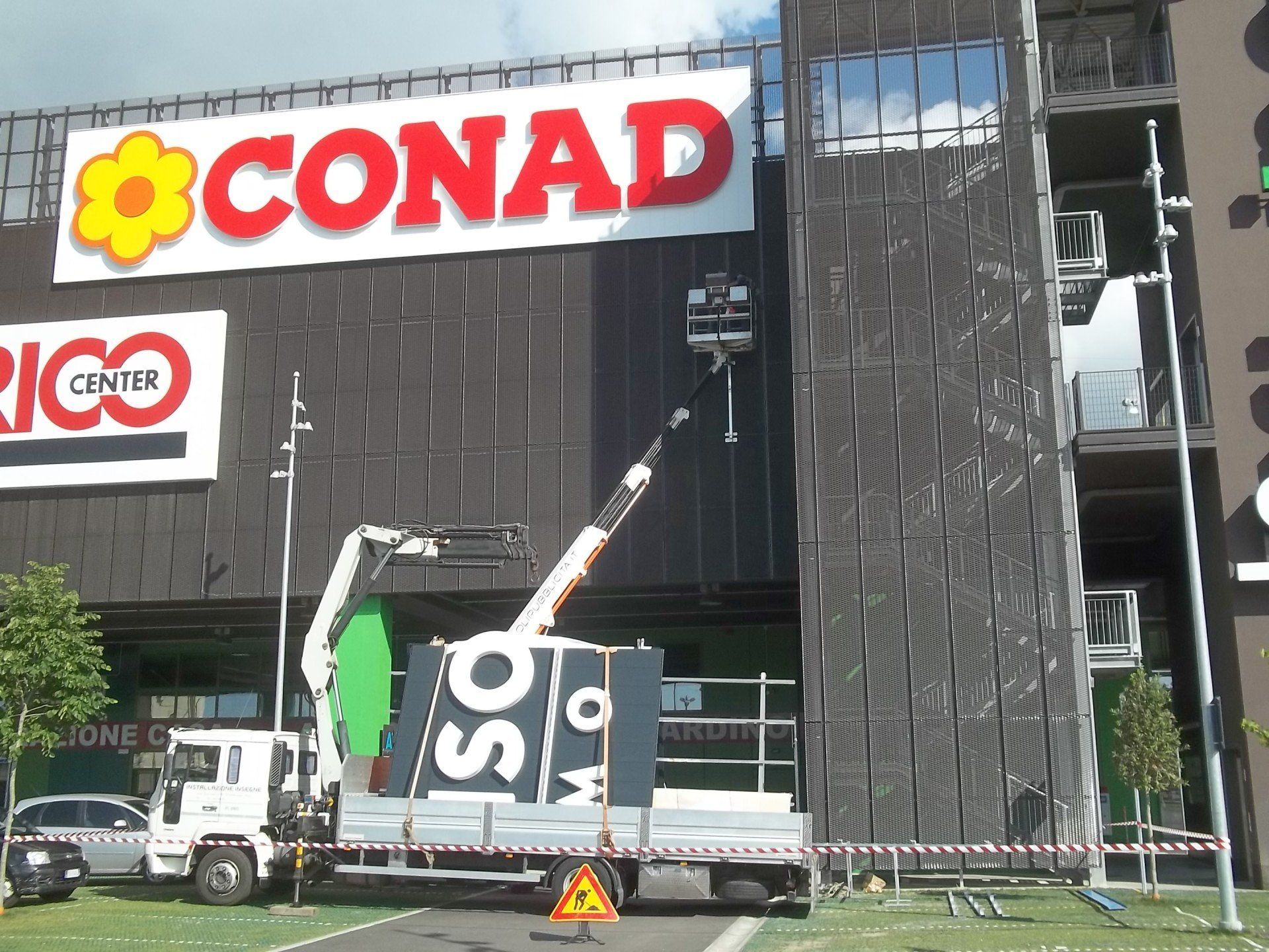 Un camion con gru installa una nuova insegna per un punto vendita SO...?, all'esterno di un supermercato CONAD.
