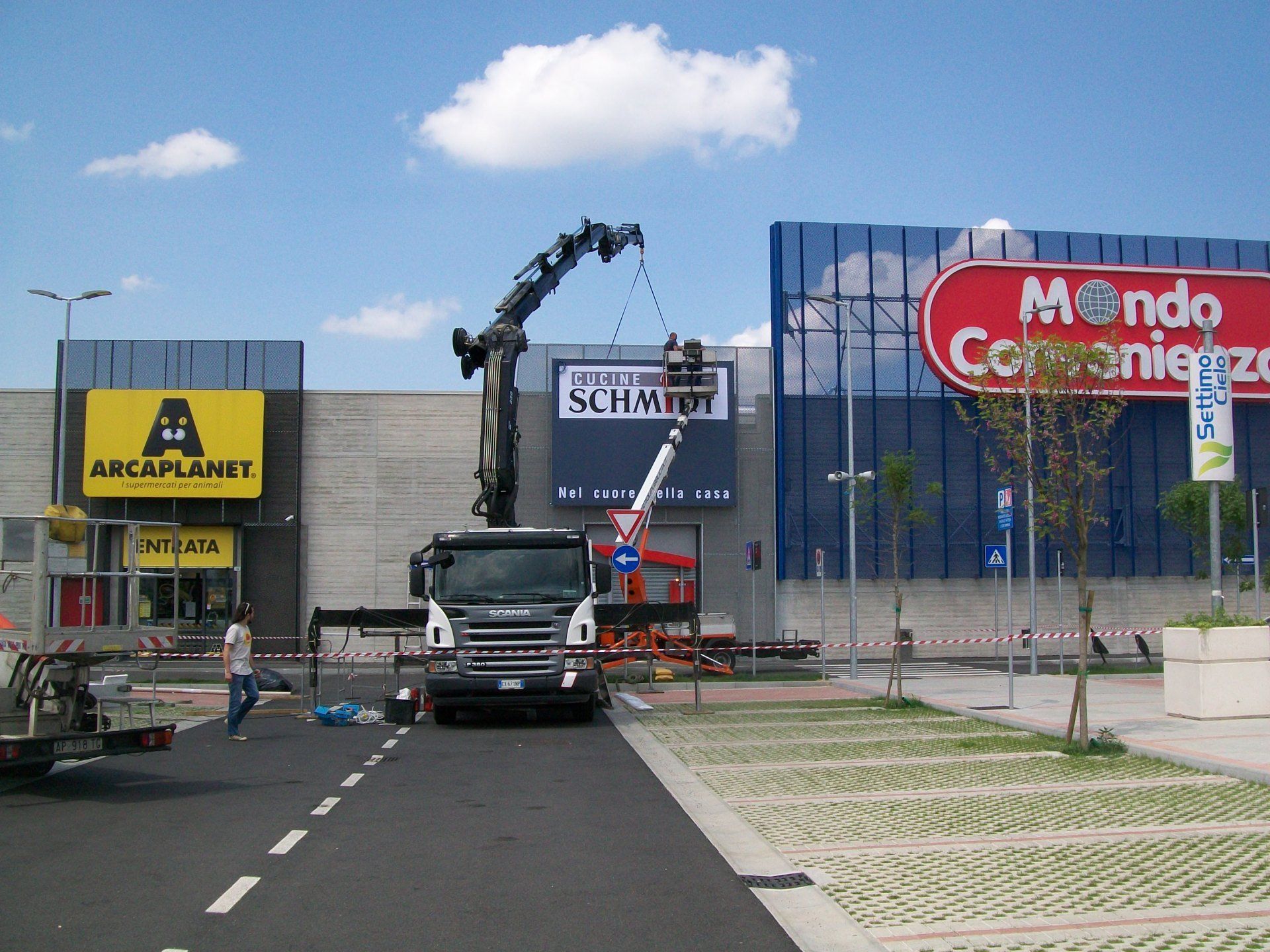 Gru montata su camion che installa un'insegna in un centro commerciale. Gli operai sono presenti; il cielo è azzurro.