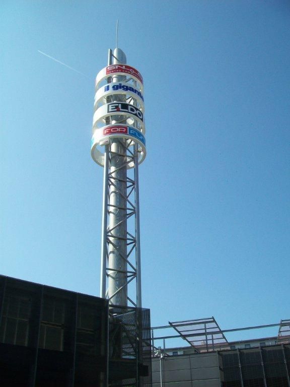 Alta torre cilindrica con loghi pubblicitari sullo sfondo del cielo azzurro.