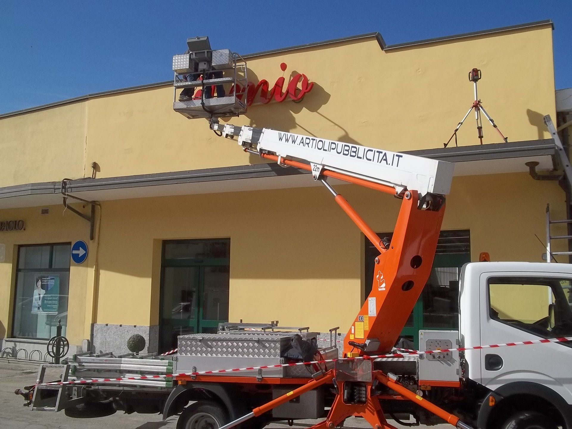 Un sollevatore montato su un camion si protende verso l'insegna di un negozio; presumibilmente un operaio lo sta riparando.