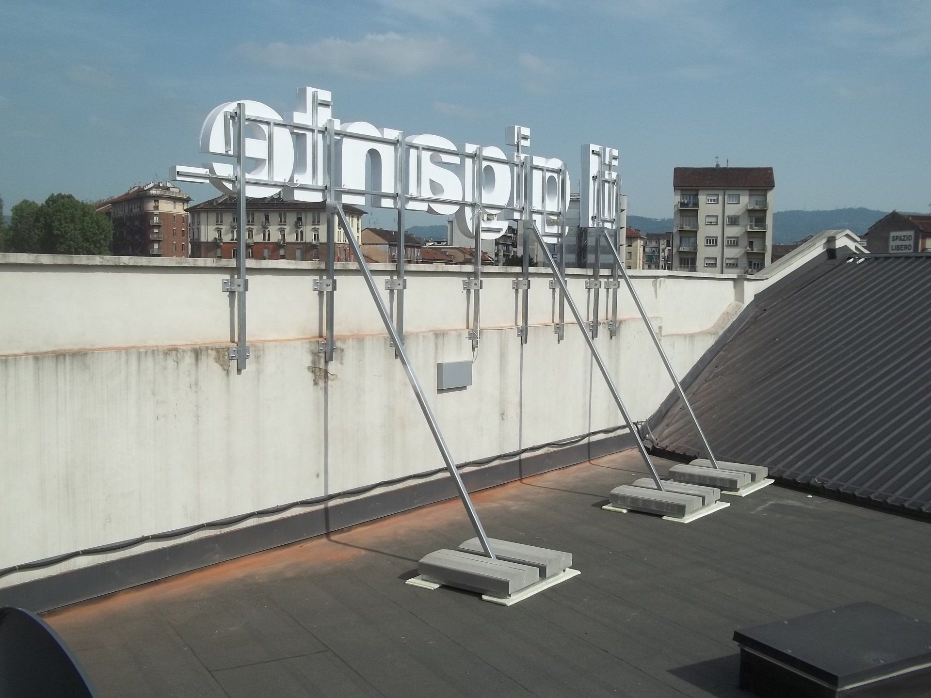 Segnaletica sul tetto di un edificio, sostenuta da rinforzi metallici e basi in cemento, con edifici sullo sfondo.
