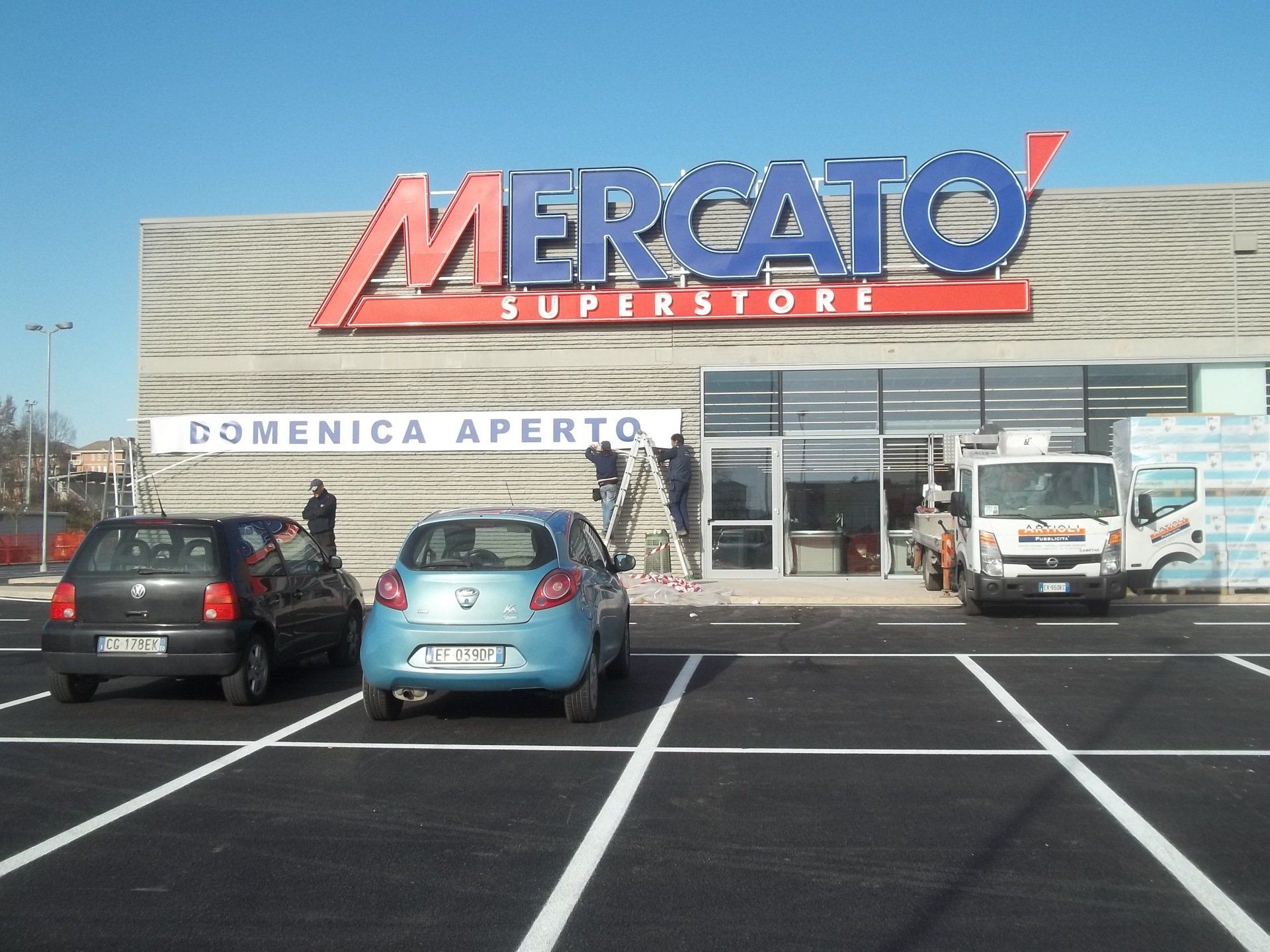 Esterno del supermercato Mercato con auto parcheggiate, operai e cartello 