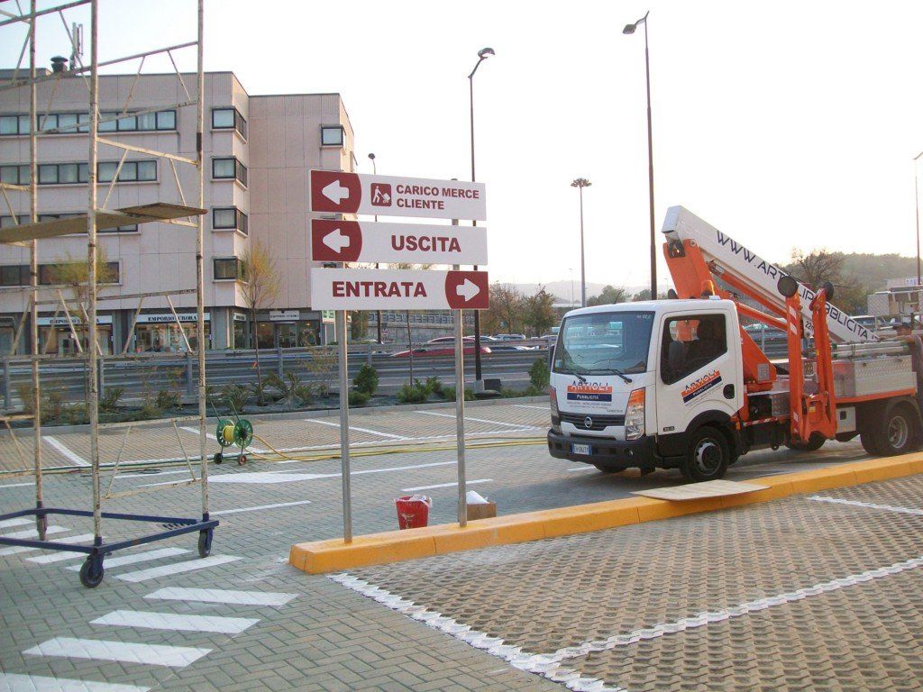 Segnaletica di ingresso e uscita di fronte a un parcheggio con un camion dotato di sollevatore.