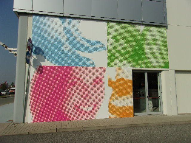 Murale colorato in stile pop art sulla facciata di un edificio, raffigurante volti sorridenti in blu, verde, rosa e arancione.
