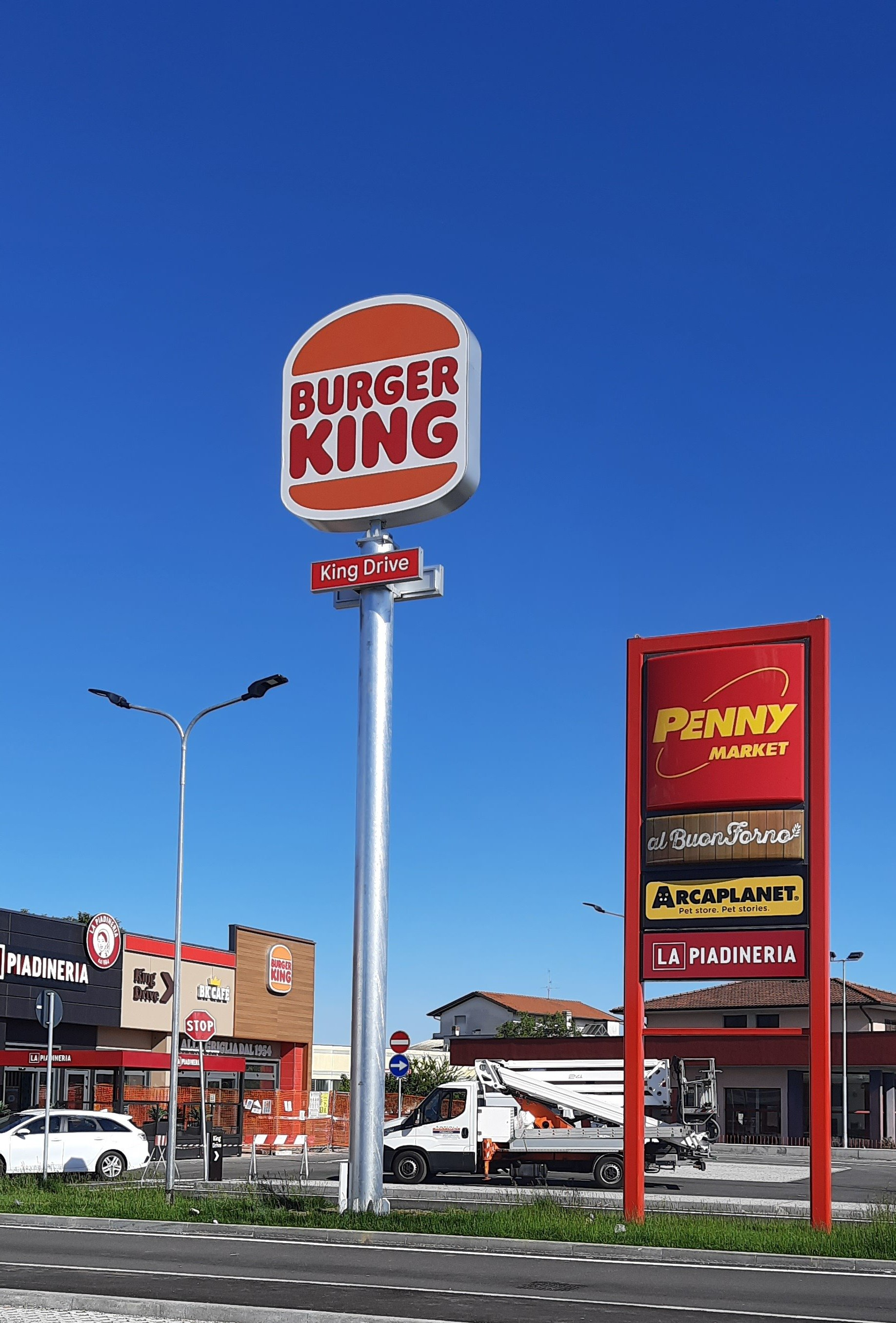 Insegna di Burger King con logo rosso e arancione, accanto a un centro commerciale con un cielo azzurro e limpido.