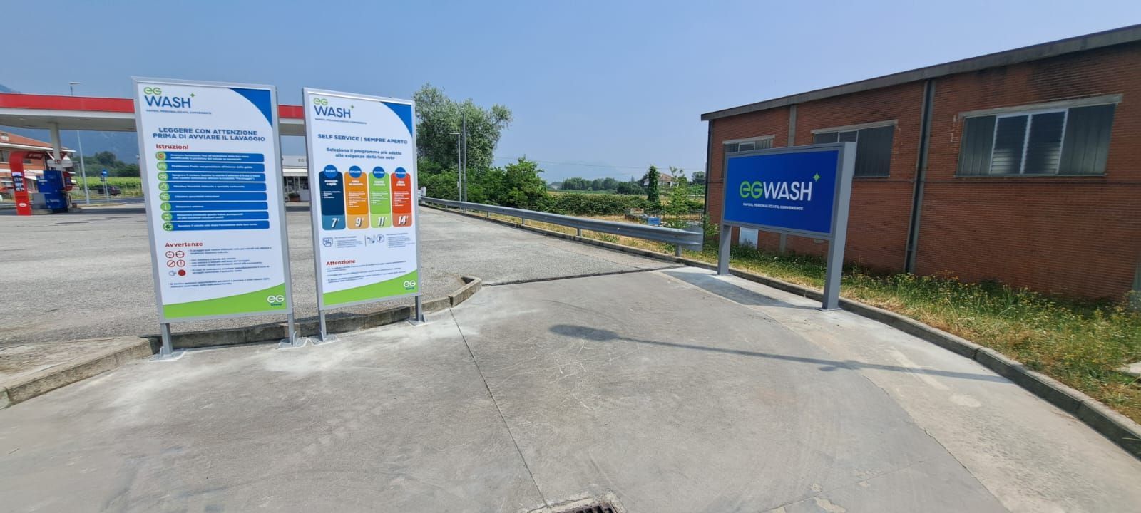 Ingresso di un autolavaggio con cartelli sulla sinistra e un edificio con un cartello sulla destra. Giornata di sole.