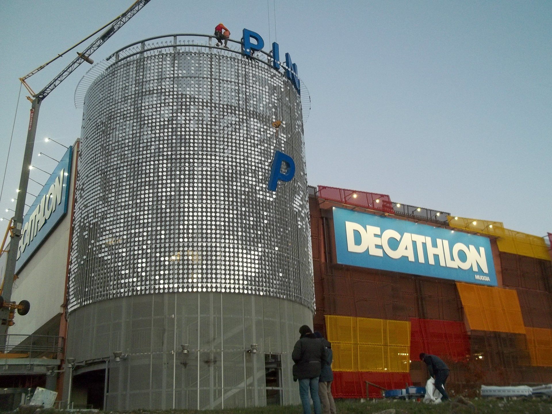 Un negozio Decathlon in costruzione con una torre argentata e una gru, cielo azzurro.