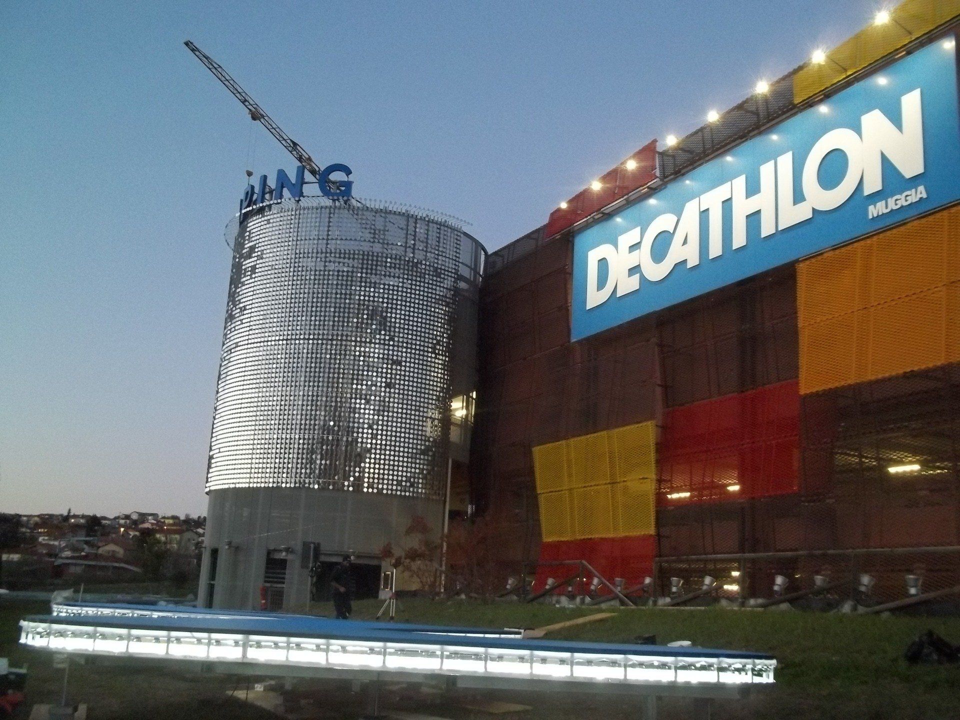 Esterno di un negozio Decathlon con una grande insegna. È in costruzione una struttura cilindrica con la scritta 