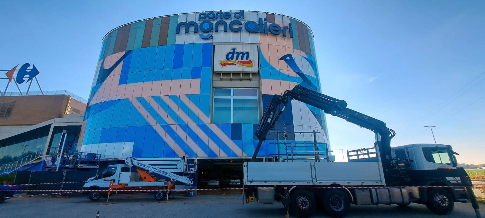Un camion con una gru accanto a un centro commerciale, con un cartello Carrefour e un negozio DM.