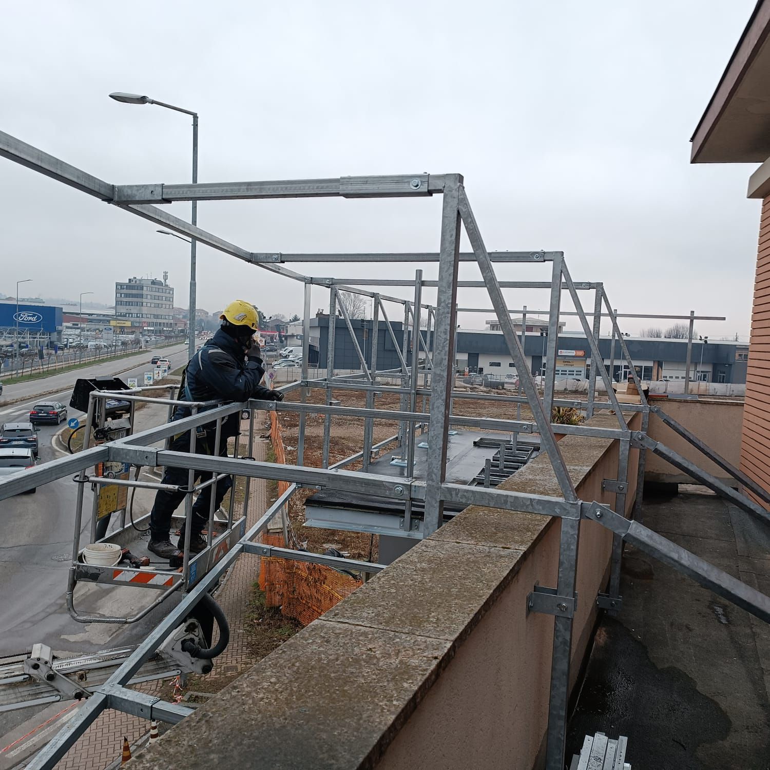 Operaio edile in ascensore, mentre installa una struttura metallica sul tetto di un edificio. Sullo sfondo la città con il cielo coperto.