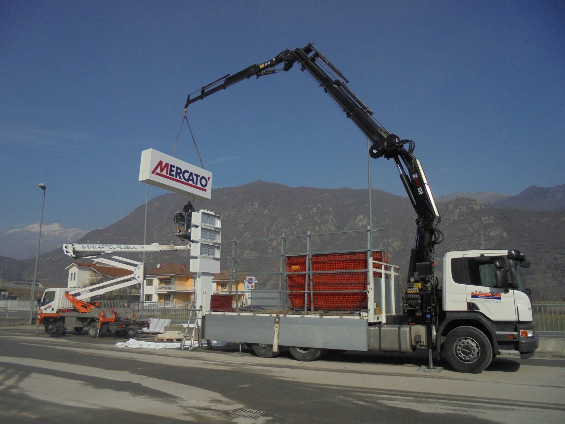 Un camion con gru solleva un cartello rettangolare con la scritta 