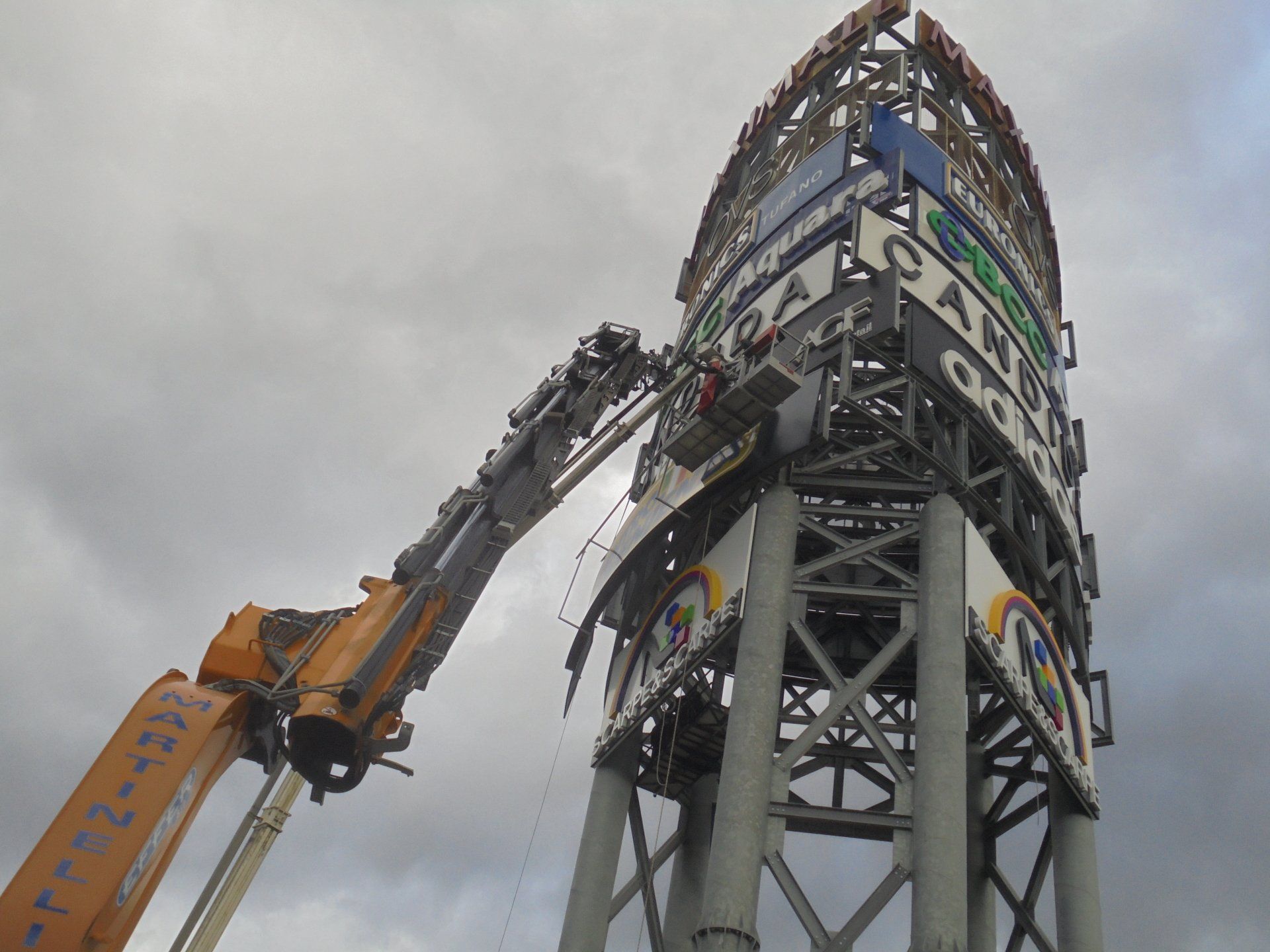 Gru che rimuove un grande cartello; parzialmente demolito con struttura metallica, cielo coperto sullo sfondo.