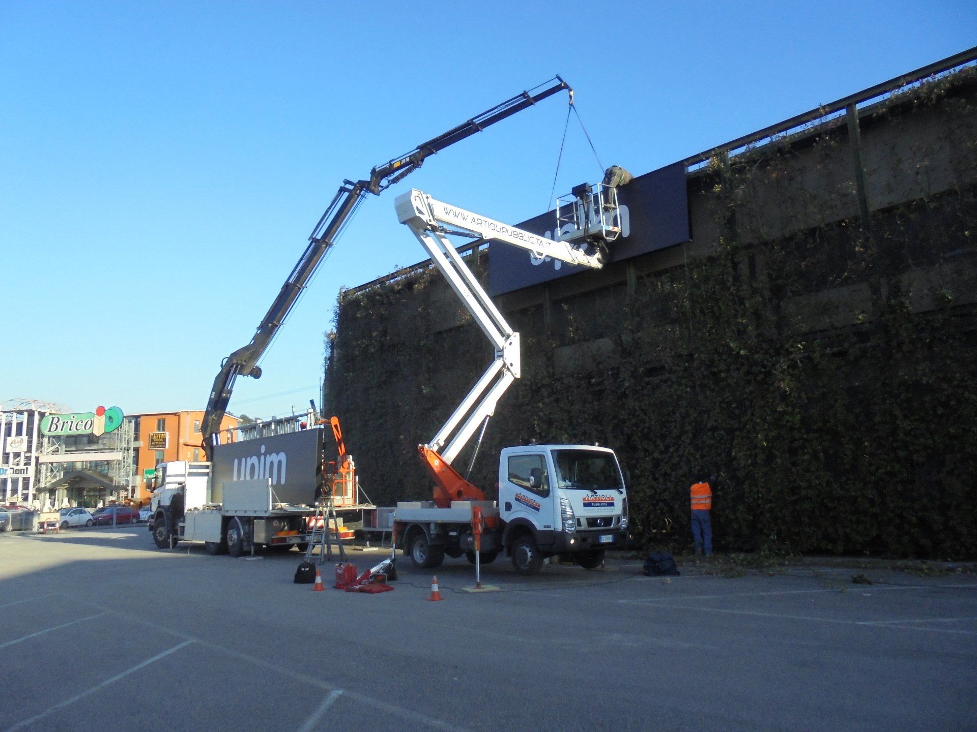 Due camion con gru stanno installando un cartello scuro su un muro ricoperto di tralci verdi; una persona con un giubbotto arancione è in piedi accanto.