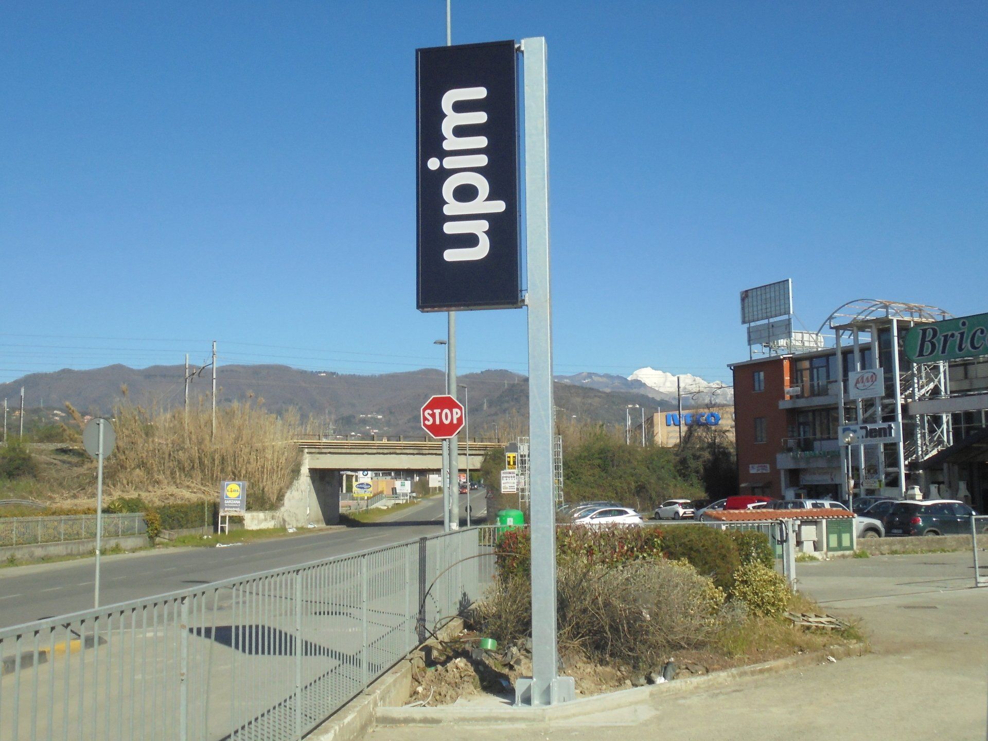Insegna per i grandi magazzini Upim. Rettangolo nero con testo bianco su un palo di metallo, strada di fronte.