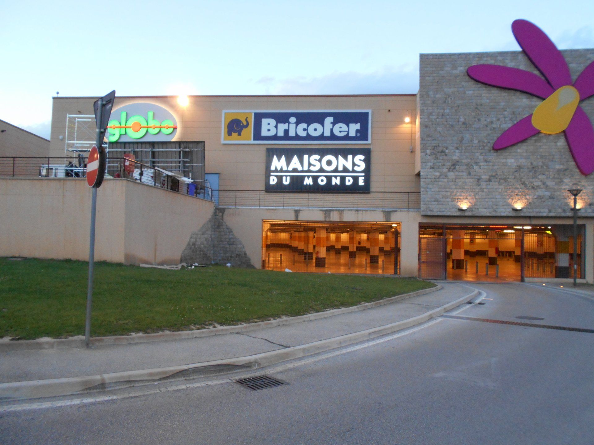 Vista esterna di un centro commerciale con insegne dei marchi Globi, Bricofor e Maisons du Monde. L'edificio è decorato con grandi fiori.